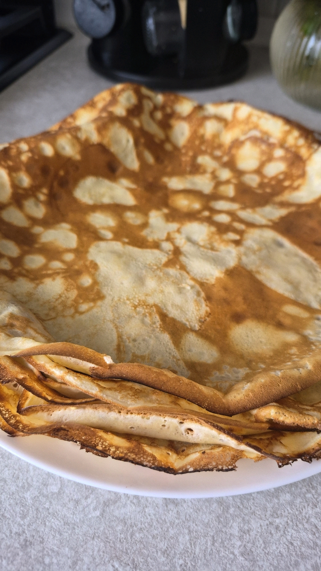A close-up, overhead shot shows a stack of crepes on a white plate. The crepes are golden brown and have a slightly uneven texture with some darker, crispier edges. The plate is set on a textured, light-colored surface, possibly a countertop. In the background, slightly out of focus, is a black kitchen appliance and a glass vase with a green sprig. The scene depicts a domestic setting, likely a kitchen, and is not specific to any particular city or location within Krauja, Latvia.