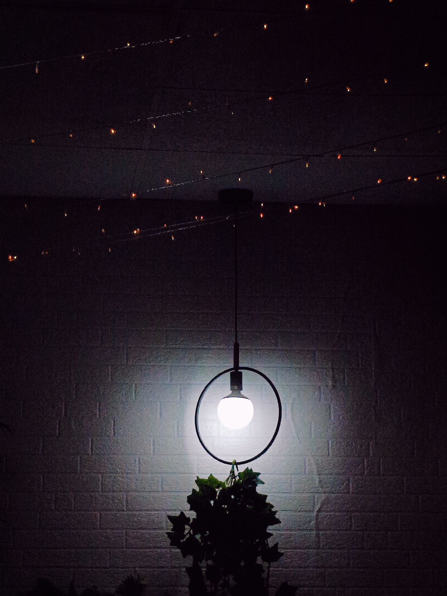 The image depicts an indoor scene, likely a room, with a prominent hanging lamp. The lamp features a circular metallic frame enclosing a bright, illuminated bulb, suspended from the ceiling by a dark cord. Below the lamp, a silhouette of dark green foliage is visible, possibly from a potted plant. The background consists of a textured, white brick wall, illuminated by the light from the hanging lamp, which casts a soft glow and a shadow on the wall. Across the ceiling, a string of small, warm-toned lights is draped, creating a festive or ambient atmosphere. The overall lighting is dim, with the hanging lamp and string lights being the primary sources of illumination, suggesting the photograph was taken at night or in a dimly lit environment. There are no people or animals visible in the image. The provided information about the location in Rafelbunyol, Spain, is not visually evident from the image itself.