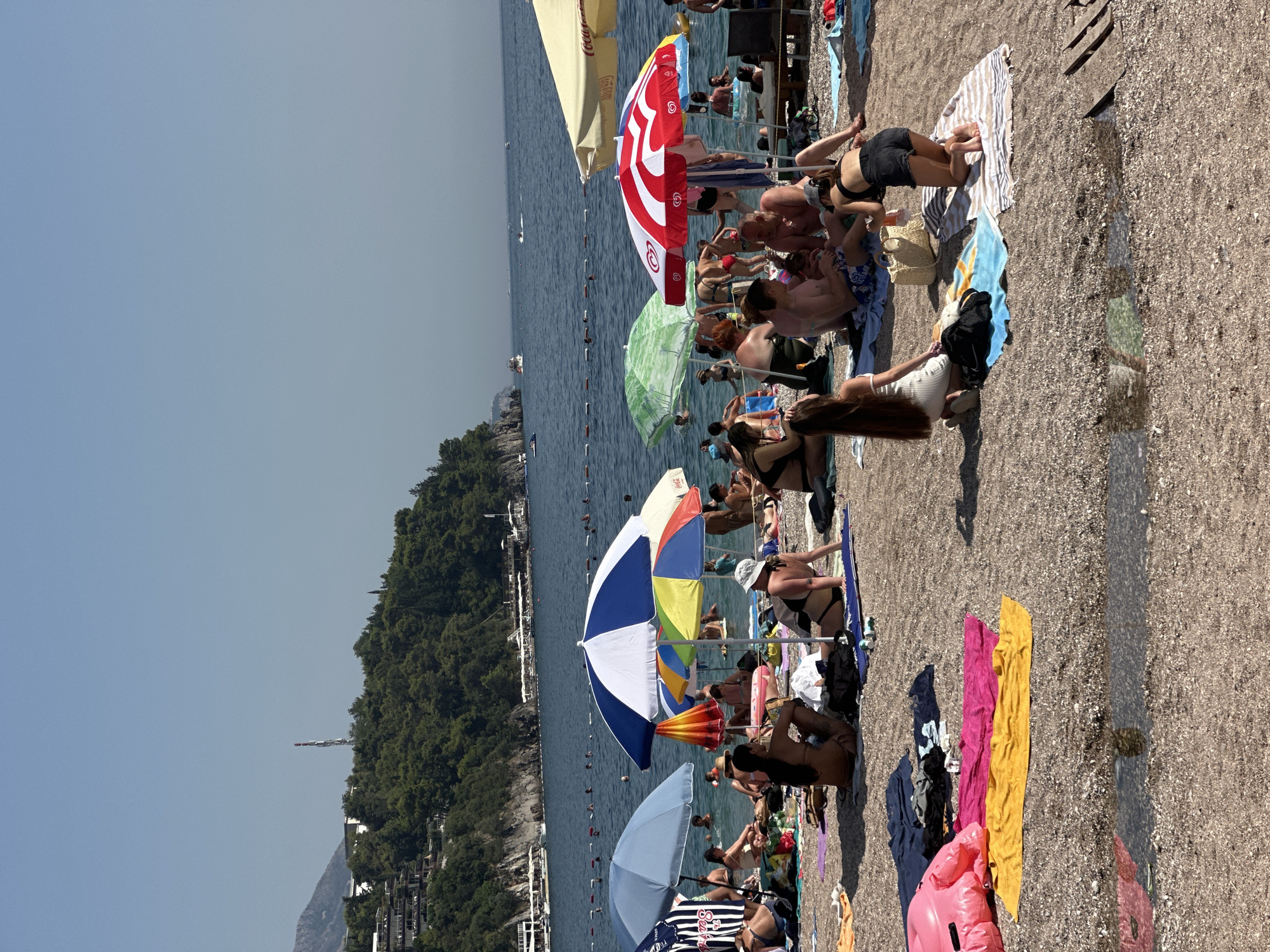 The image captures a crowded beach in Budva, Montenegro, on a sunny day. The scene depicts a variety of people relaxing on the shore and swimming in the sea. Beach umbrellas in various colors, including blue, red, and yellow, provide shade for the sunbathers. Several individuals are seated or lying on towels on the pebbled beach, while others are in the water, some swimming and others wading. The background features a lush green hillside dotted with buildings, and the calm, blue sea stretches towards the horizon. The overall atmosphere is one of a typical summer beach day, with many people enjoying the coastal environment.