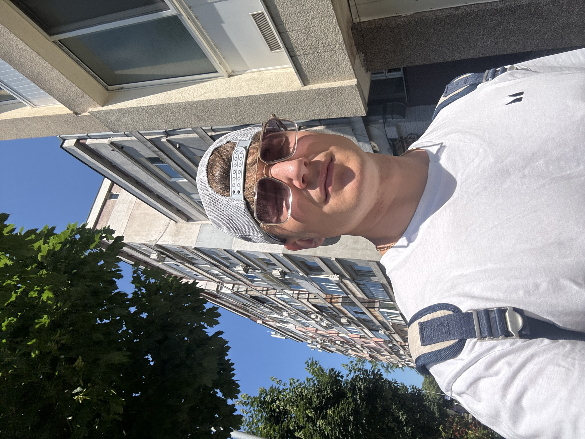 A young man wearing a white t-shirt, sunglasses, and a grey baseball cap is standing on a sidewalk in front of a tall apartment building. He is also wearing a backpack with blue and beige straps. The building behind him is several stories high with many windows. To the left of the building is a tree with green leaves. The sky is clear and blue. This scene is located on ul. "Gen. Skobelev" street in Burgas, Bulgaria.