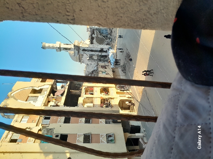 This image depicts an outdoor scene in Khan Yunis, Palestinian Territory, viewed from an elevated perspective through vertical metal bars. In the immediate foreground, a quilted white fabric is visible on the bottom left, and a dark, indistinct object partially obscures the bottom right.
On the left side of the frame, a multi-story building with a light-colored facade exhibits significant structural damage, including partially collapsed sections, exposed interiors across multiple floors, and numerous broken or missing windows. Arched architectural details are visible on its upper levels.
In the mid-ground, a paved street with white lane markings stretches horizontally. The street is bordered by piles of rubble and debris, particularly on the right side.
Further in the background, to the right, a mosque featuring a prominent minaret and a domed roof stands, also showing substantial damage to its structure and surrounding grounds.
Several individuals are present on the street: two men are walking together in the mid-ground, a group of people is gathered near the damaged mosque, and other individuals are visible on the left sidewalk and walking away in the bottom right corner of the street. The sky above is clear and blue.