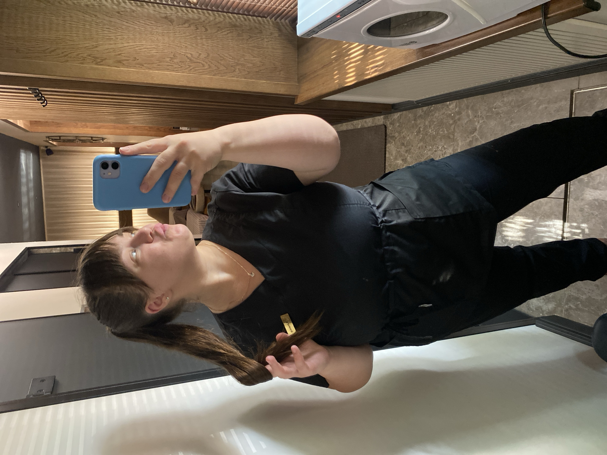 A Caucasian woman with brown hair in a ponytail and bangs, wearing a black V-neck scrub top and black scrub pants, is taking a mirror selfie in what appears to be a spa or salon in Votkinsk, Russia. She is holding a blue iPhone in her right hand and has her left hand in her hair. She is puckering her lips. A golden name tag is visible on her left chest. The background shows a wooden paneled wall with hooks, a partially visible beige curtain, and a window with blinds. A white machine with a circular opening is on a wooden counter to the right. The flooring is a marble-patterned tile.