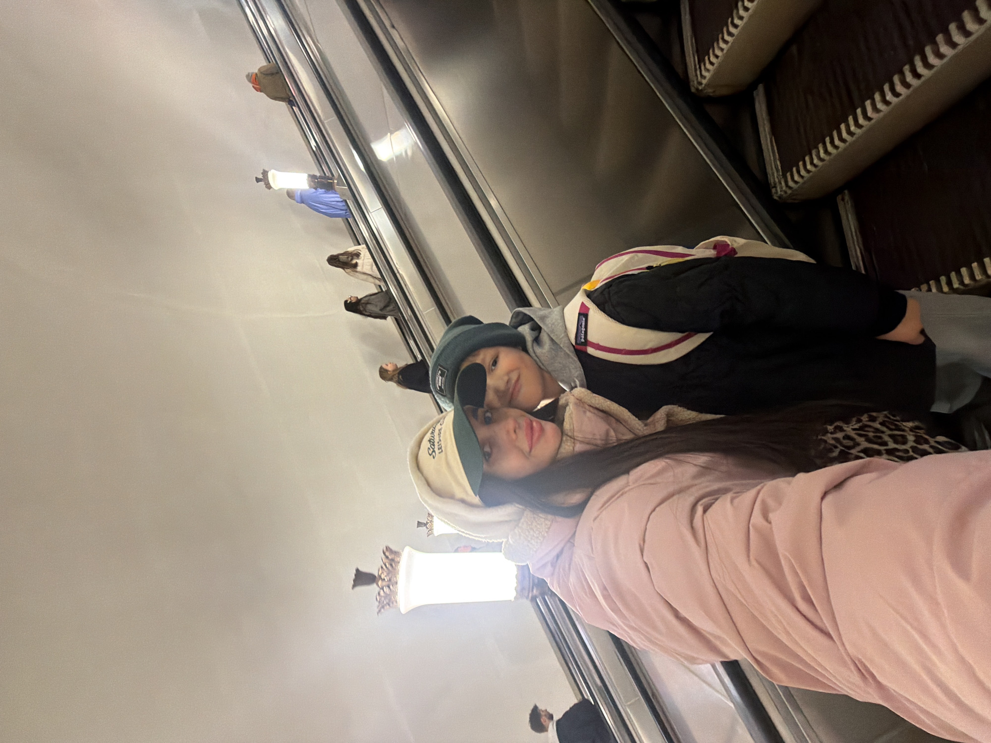 A selfie-style photograph captures two individuals on an escalator within the Saint Petersburg Metro system in Saint Petersburg, Russia. In the foreground, a female adult with long dark hair, wearing a pink puffer jacket, a cream-colored hood, and a beige and green baseball cap with "Saturday LEISURE C" text, directly faces the camera with a slight smile and visible pink lip makeup. To her right and slightly behind, a child, likely male, wears a dark beanie, a gray hooded sweatshirt under a black jacket, and a white backpack with pink stripes and a "Patagonia" logo; he also looks towards the camera. Both individuals are positioned on a wide, ascending escalator with metallic handrails and dark steps with serrated edges.
In the mid-ground, a prominent ornate lamppost with a frosted globe and decorative bronzed top stands between ascending and descending escalators. Further up the ascending escalator, multiple indistinct figures are visible: one in a dark jacket with glasses, another in a light-colored jacket, a third in a bright blue jacket, and a fourth in a dark jacket with an orange scarf or hood. On a parallel descending escalator to the far left, a male figure in a dark jacket with a white hood is also visible. The background consists of plain, light-colored walls and a ceiling, illuminated by the escalator lighting and the ornate lampposts, indicative of a deep underground metro station.