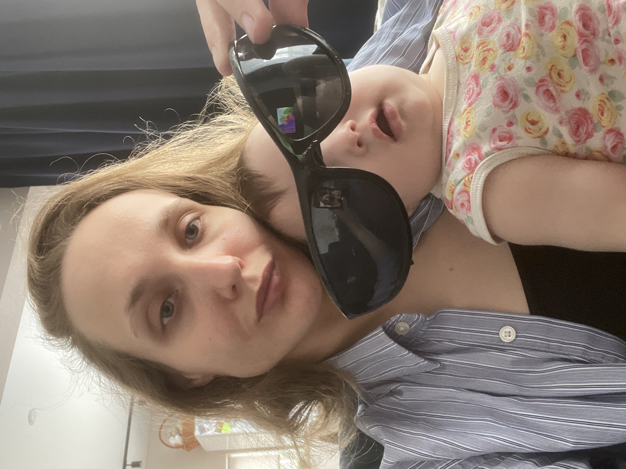 A woman with blonde hair is holding sunglasses in front of her face, with a baby wearing a floral patterned top lying on her chest. The woman is wearing a blue striped shirt. The sunglasses reflect an image. The location is unknown and is not confirmed to be Подшивалово, Russia.