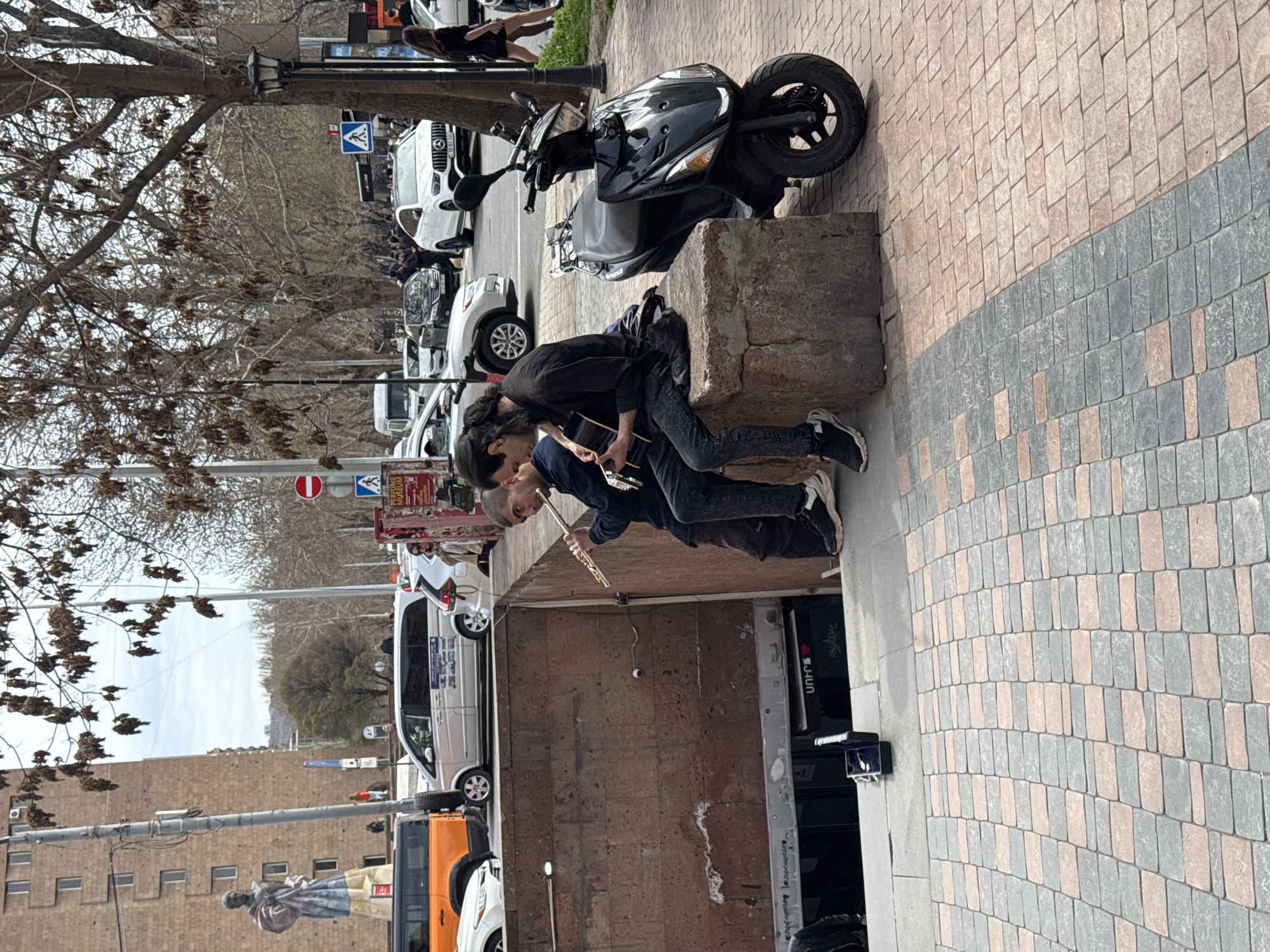 Two men are playing music on a city street. One man is playing a guitar, and the other is playing a flute. They are sitting on a concrete block. A black scooter is parked near them. In the background, there is a line of cars and trees. The photograph was taken on a street in Yerevan, Armenia. A statue and a sign for the Perfume Museum are visible in the background.