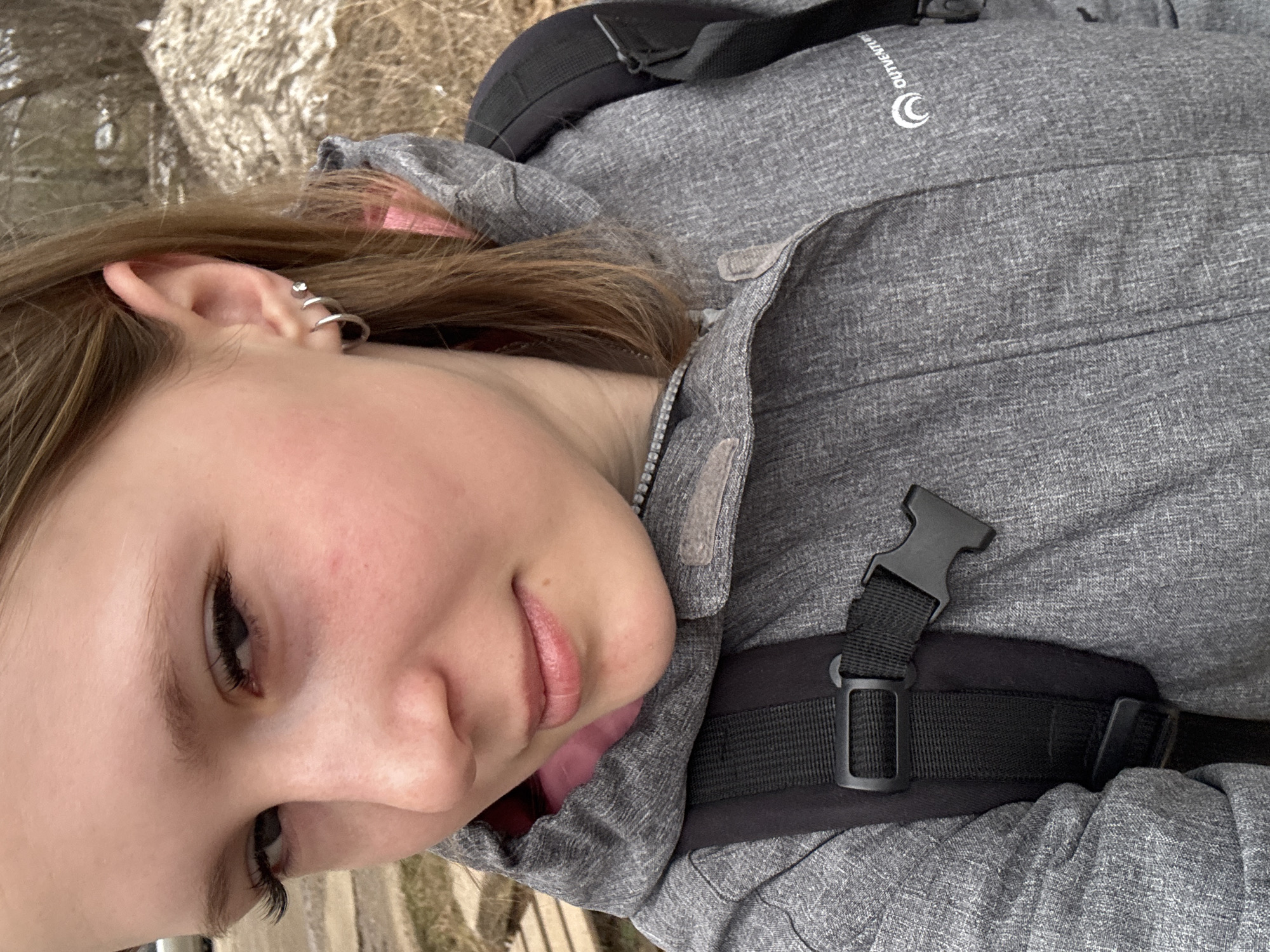 A young woman is pictured wearing a grey jacket and backpack. Her left ear is visible, with multiple earrings. The jacket's brand logo "Outventure" is visible. A black backpack strap with a buckle crosses over her torso. The background appears to be natural and blurry, suggesting an outdoor setting. The image location cannot be determined to be Chaikovsky, Russia.