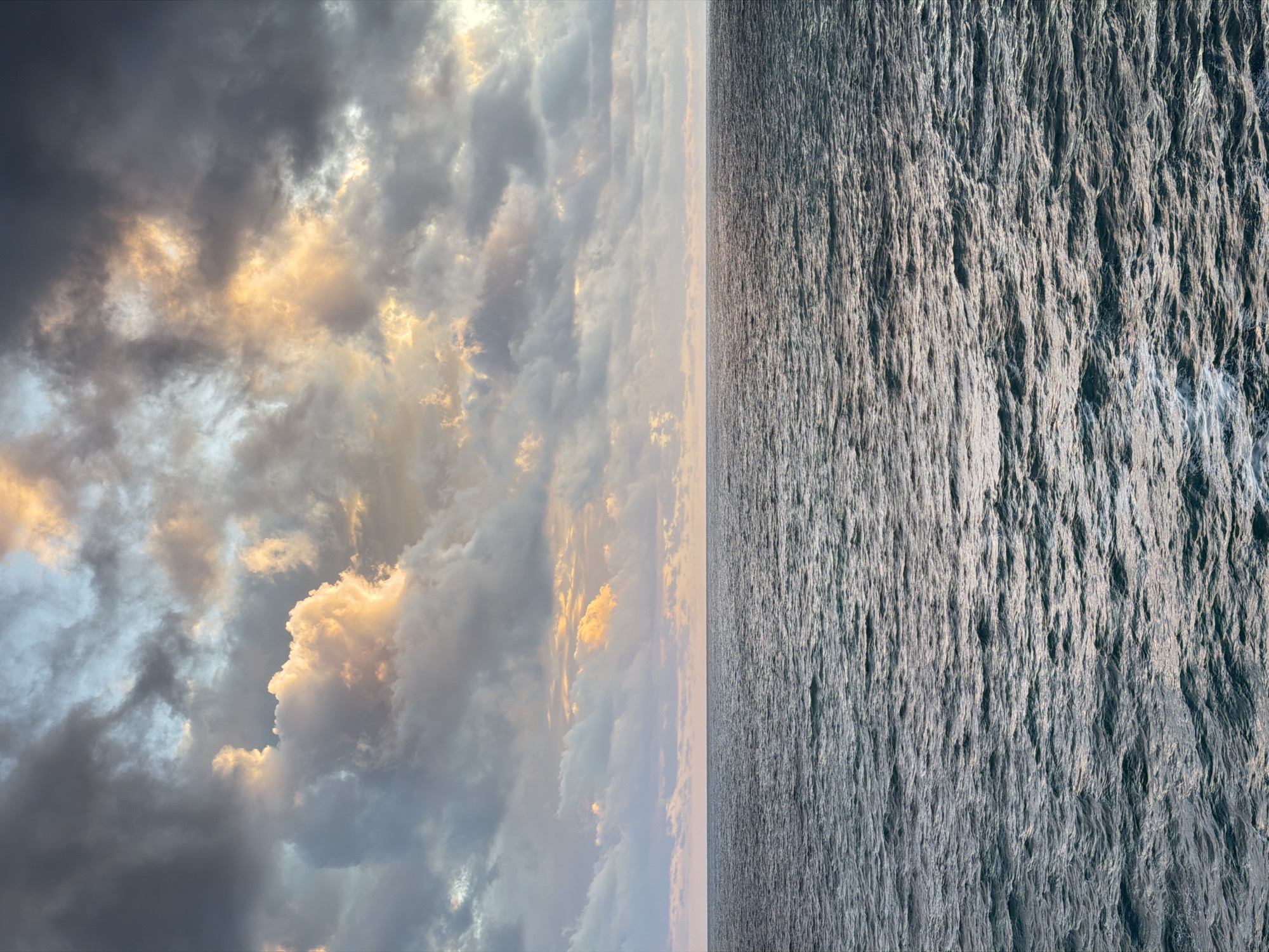 The image depicts a scene of an ocean view from Petrovac centar, Montenegro. The left side of the image features a sky filled with dark, stormy clouds, some illuminated with golden light breaking through. The right side shows the ocean, with its surface rippled and textured by wind. The photograph is oriented vertically, with the horizon line running horizontally through the center.