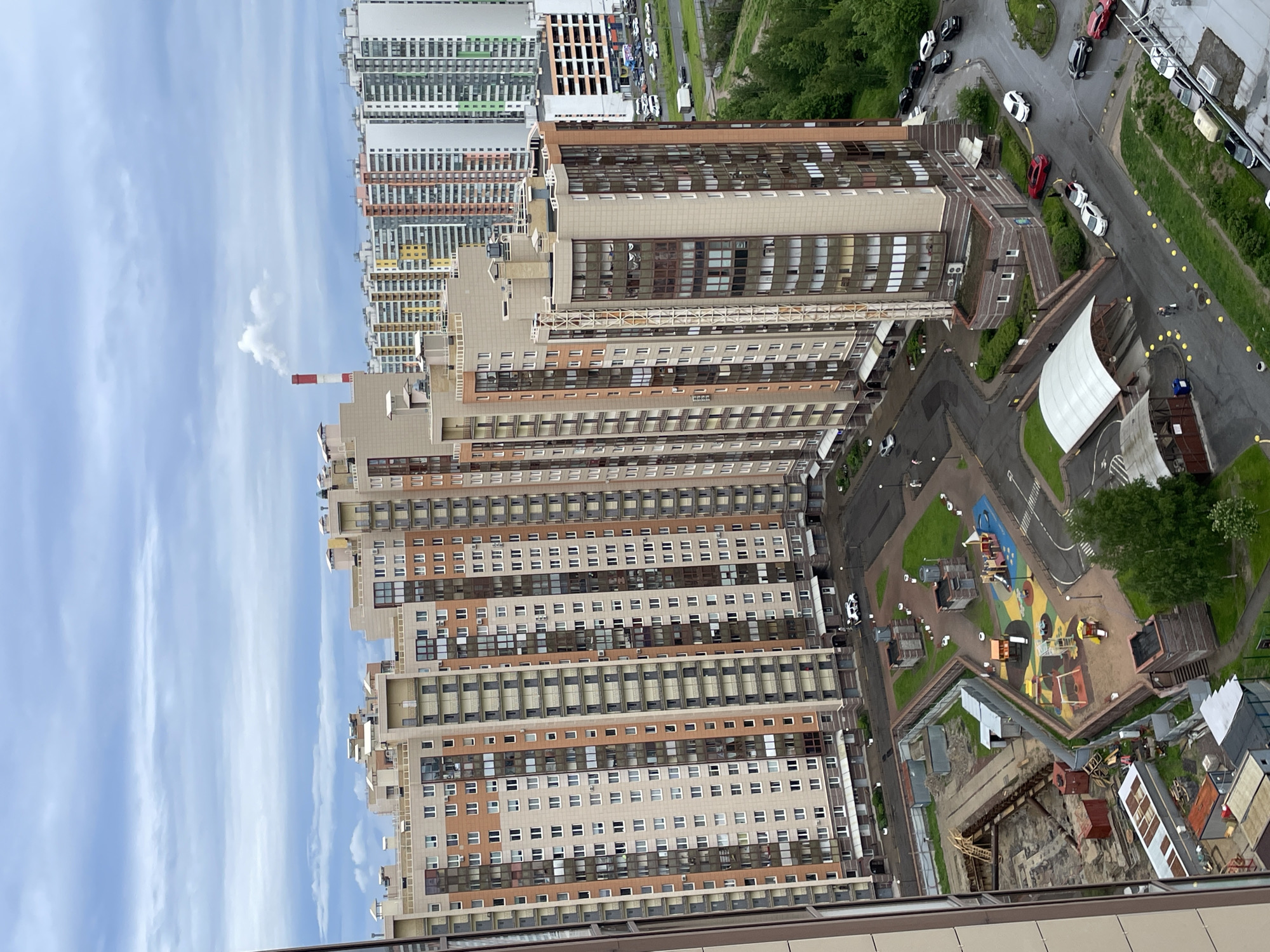 The image presents an aerial view of an urban landscape, likely in Saint Petersburg, Russia, characterized by a mix of high-rise residential buildings and green spaces. Several multi-story apartment complexes are visible, displaying a variety of architectural styles and colors. The buildings are primarily beige and brown with repeated window patterns, alongside taller, white and colorful structures in the distance. A smokestack emitting a plume of smoke is present on one of the buildings. Below the buildings, there's a playground with colorful equipment and design on the grounds, indicating an area for recreational activities. A road network with parked and moving cars is visible, suggesting a typical urban traffic pattern. The environment is predominantly cloudy skies, with trees providing a green element in the urban setting. The scene conveys a sense of urban density mixed with elements of community and recreational space.