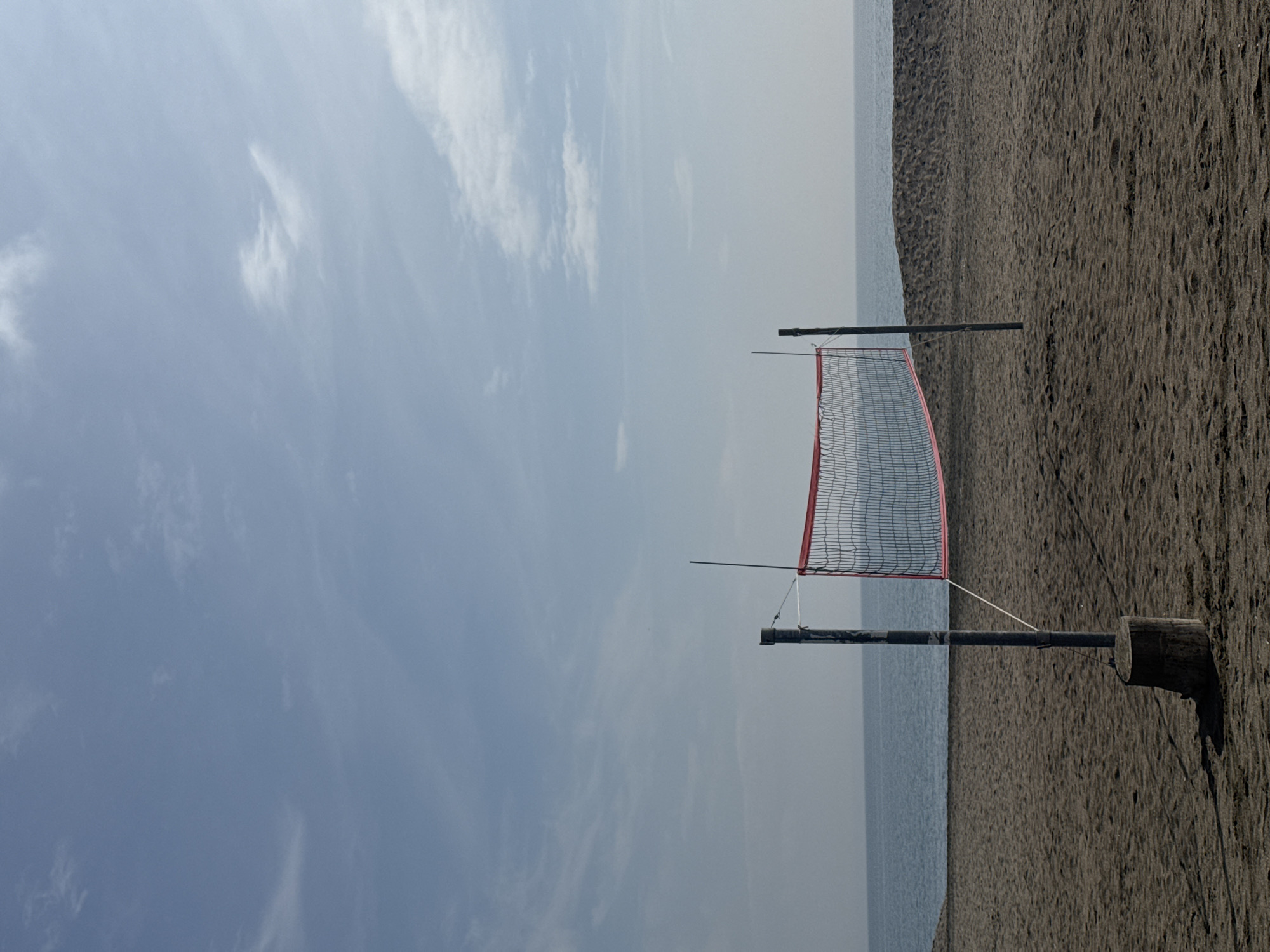 The image shows a beach volleyball net standing on dark sand in Burgas, Bulgaria. The sea is visible in the background, separating the sand from the cloudy sky. There are no people visible in the scene. The net has a red trim and the poles appear to be made of dark wood. A log is situated at the bottom of the right pole, perhaps to secure it.