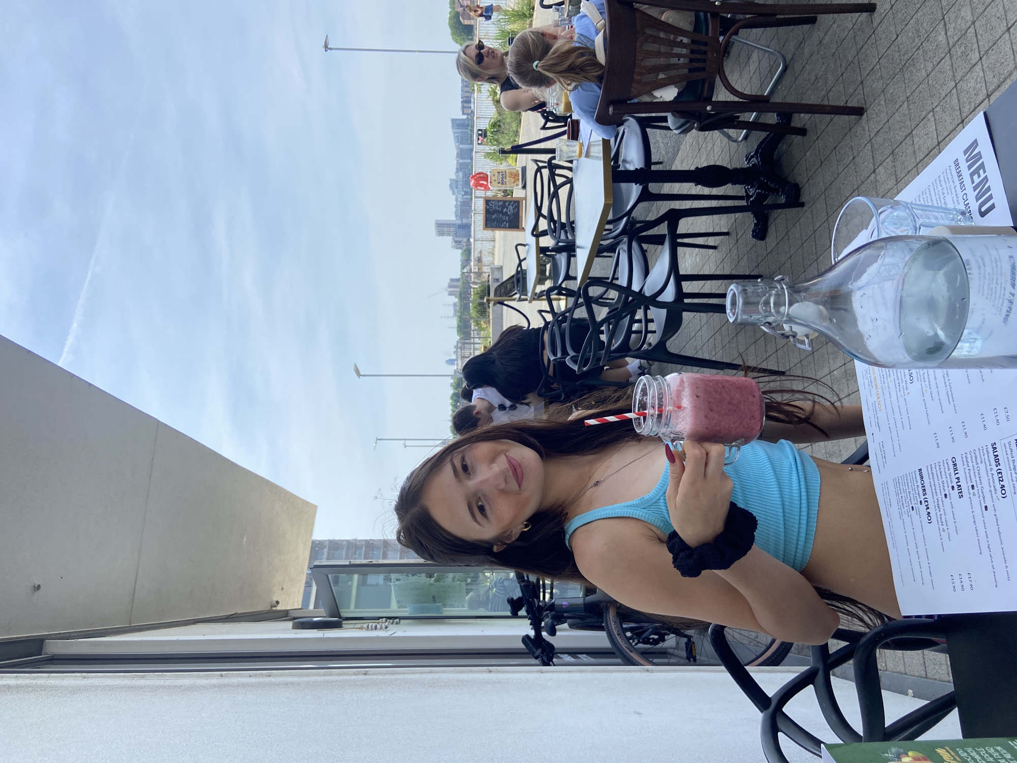 The image captures a young woman seated at an outdoor table, holding a smoothie. She has long brown hair, is wearing a light blue top, and has a hair tie on her wrist. A menu, a water bottle, and a glass are on the table. In the background are other patrons at tables, buildings, and a view of the city.
The location appears to be in London, United Kingdom, based on the visible buildings and architecture.