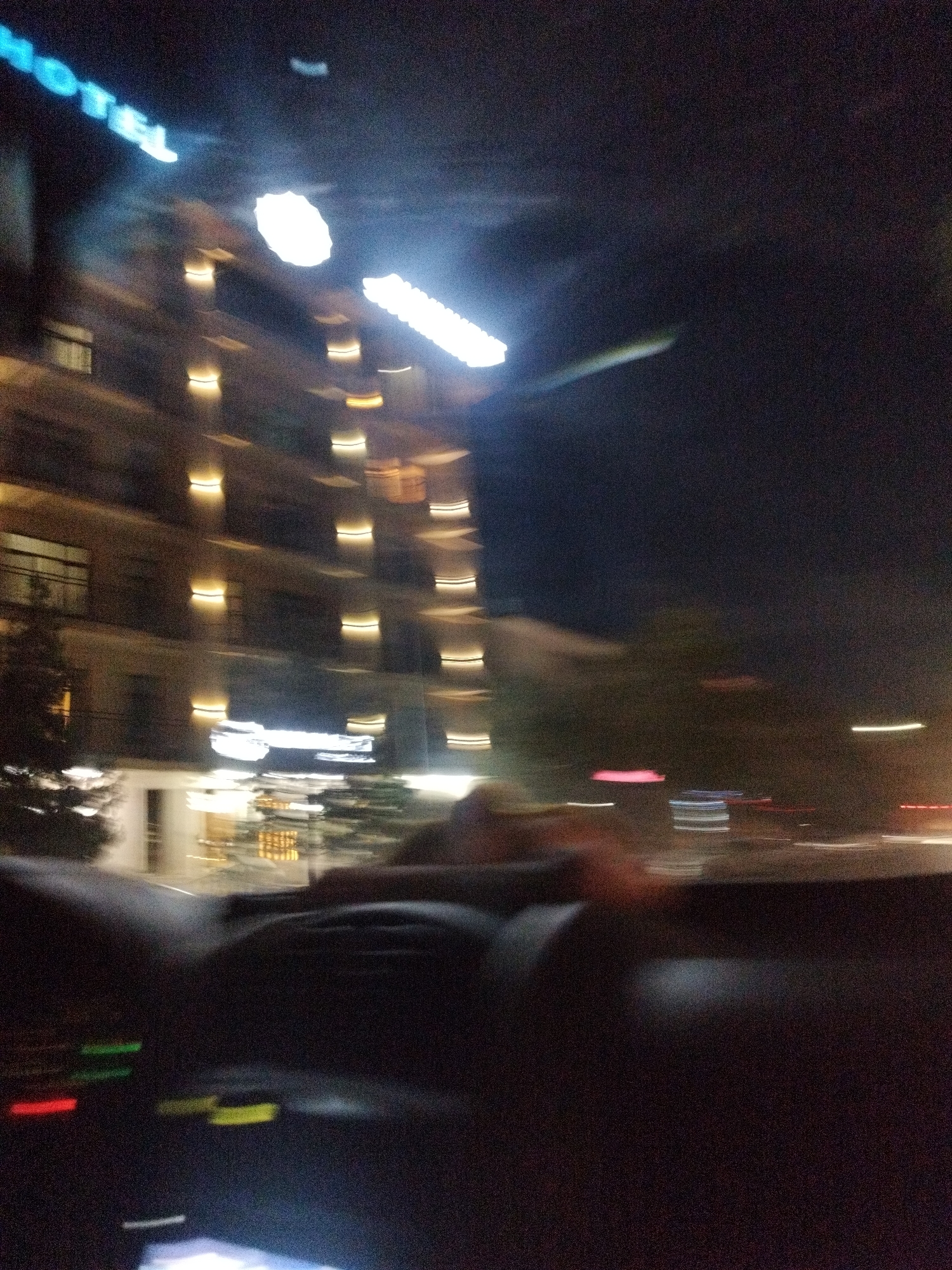 The image captures a blurry nighttime scene from inside a vehicle, likely at a standstill. In the background, a multi-story building with illuminated windows suggests a hotel, as indicated by the glowing blue "HOTEL" sign visible on the upper left. The lights from the building create a streaking effect due to motion blur. In the foreground, the blurred interior of the car is visible, with parts of the dashboard and steering wheel dimly lit. Several small, indistinct colored lights on the dashboard are also apparent. The overall impression is one of motion and a fleeting glimpse of urban nighttime activity, possibly along a street in Shymkent, Kazakhstan.