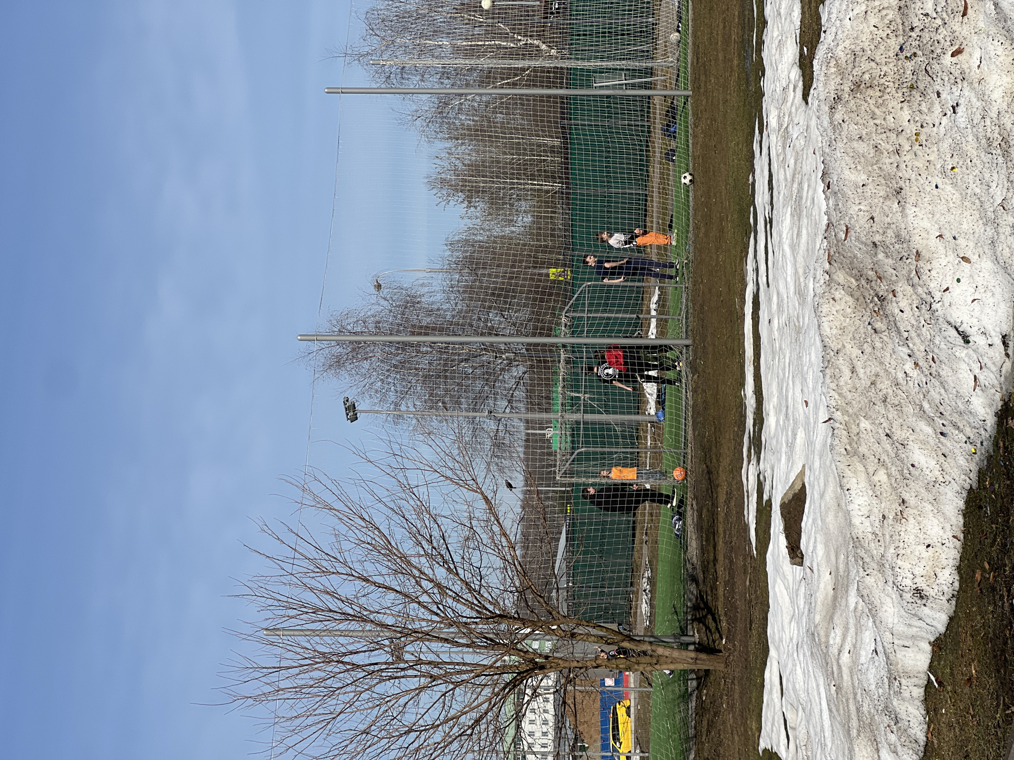 The image shows a group of people on a soccer field in Moscow, Russia. A soccer field is surrounded by a green mesh net and silver poles. A leafless tree stands in the foreground with branches that cover some of the soccer field. A patch of melting snow is on the right side of the image, next to the field. The sky is light blue. Several people are on the field, some wearing athletic attire. At least two soccer balls are on the field. A building is visible in the background.