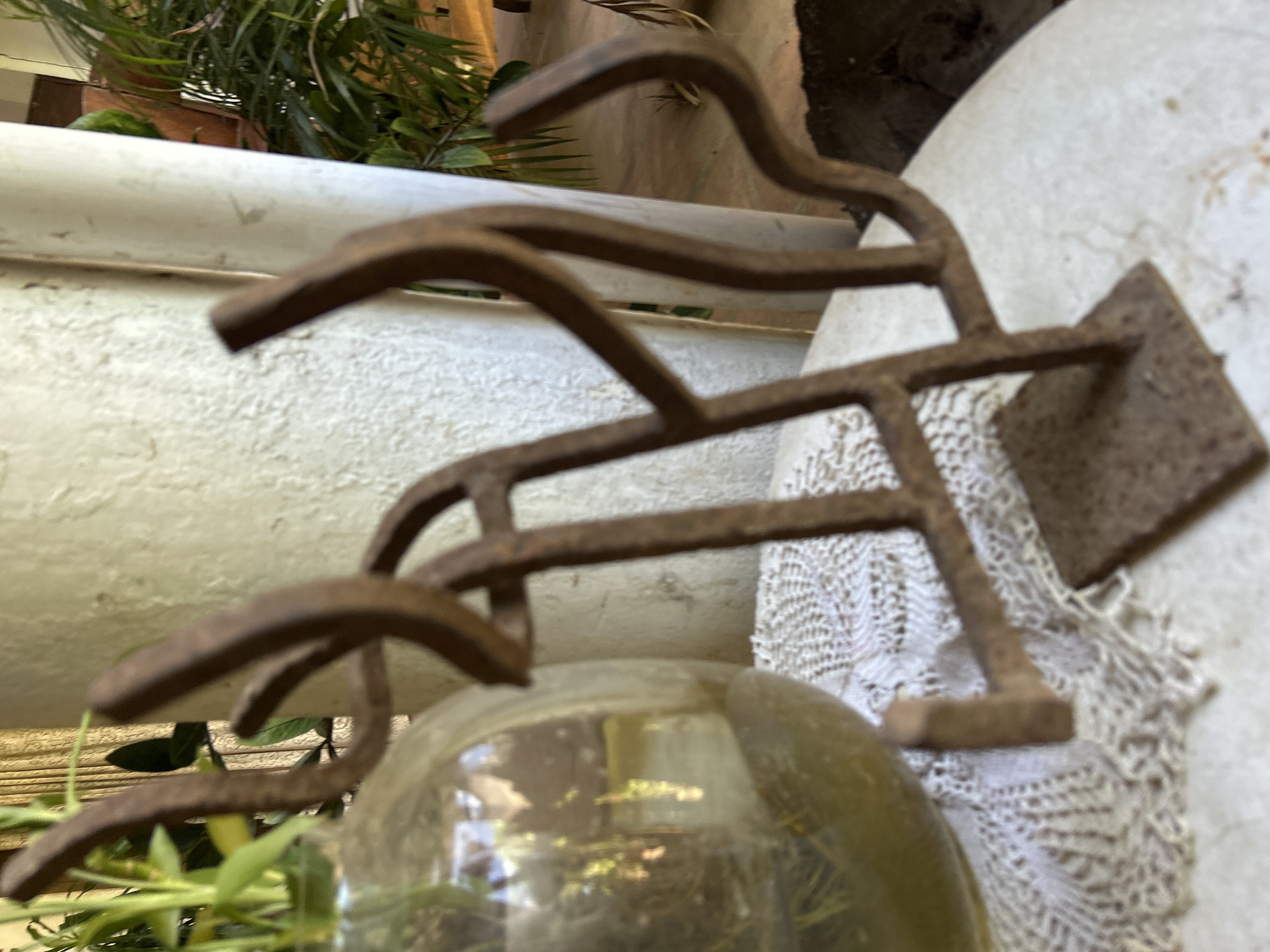 A close-up, slightly out-of-focus shot captures a weathered, rusty metal sculpture with abstract, curved elements resting on a white, textured surface. To the left, a portion of a round, glass vase with a yellowish tint contains green plant stems. The sculpture has a square base and appears to be made of bent metal rods, possibly representing an animal or an abstract form. Behind the sculpture and vase, a rough, off-white textured wall or pillar is visible, with the suggestion of green foliage and a light-colored curtain in the background. The overall lighting is soft, suggesting an indoor or shaded outdoor setting. The location is not identifiable from the image.