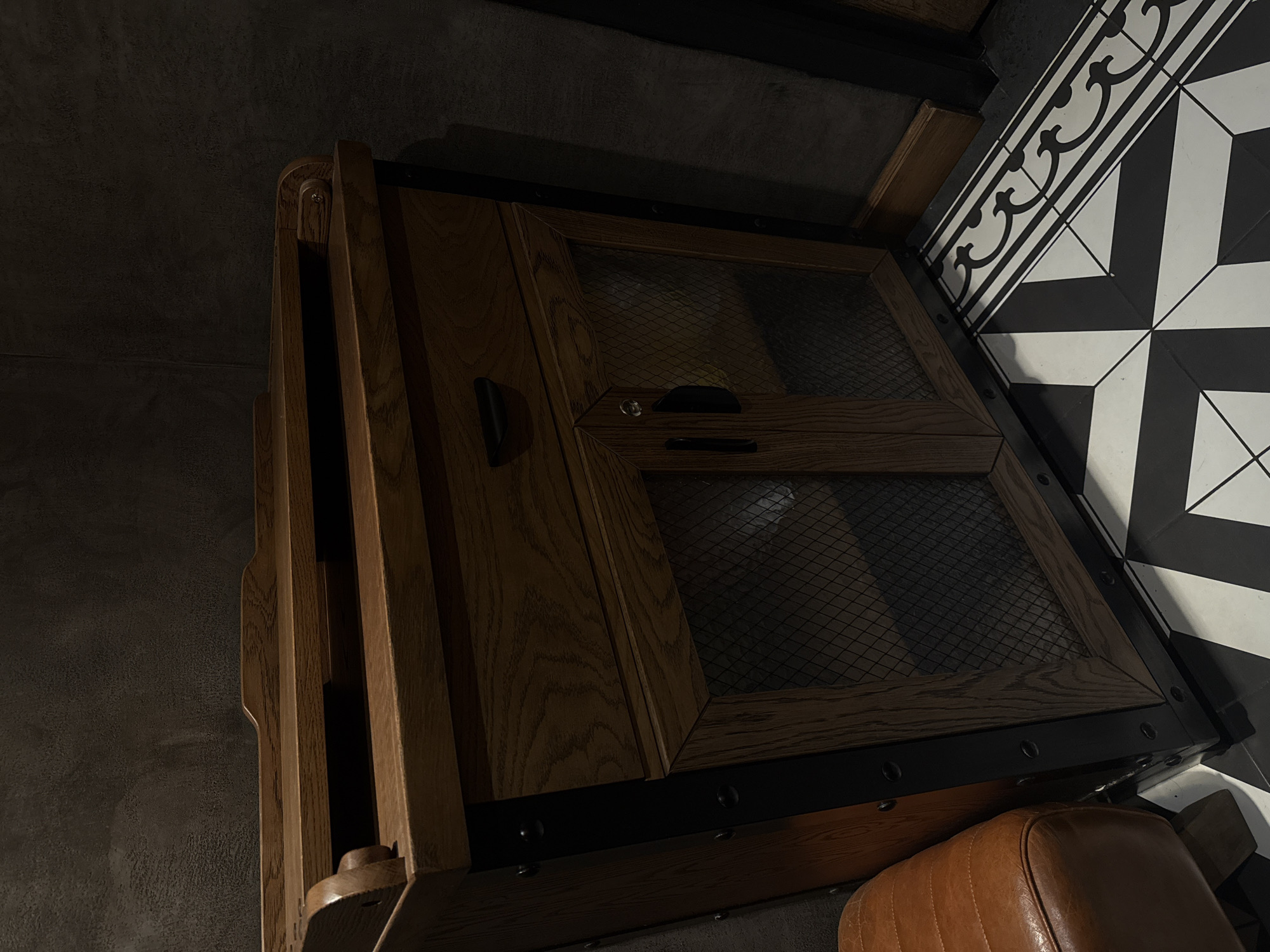 The image displays an indoor scene featuring a dark brown wooden cabinet with industrial-style accents. The cabinet's top surface includes a shallow, tray-like area formed by raised wooden edges. Below this, a single drawer with a recessed black handle is present. The lower section of the cabinet consists of two hinged doors, each framed in wood and featuring a dark, diamond-patterned mesh insert. The right door has a black handle and a keyhole. Vertical black metal straps with visible rivets or bolts run down the sides of the cabinet, enhancing its robust aesthetic.
The cabinet is positioned against a dark, textured wall with subtle variations in tone, possibly a stucco or concrete finish. The floor features a distinctive black and white tiled pattern. A border of white tiles with black scrollwork patterns runs along the base of the wall. The main floor area is covered with square tiles divided diagonally into alternating black and white triangles, forming a larger geometric pattern. To the immediate left of the cabinet, a portion of a curved, padded object upholstered in warm brown leather, exhibiting stitching and a somewhat worn appearance, is visible. The scene is dimly lit, casting deep shadows and emphasizing the textures of the materials. No individuals are present in the image.