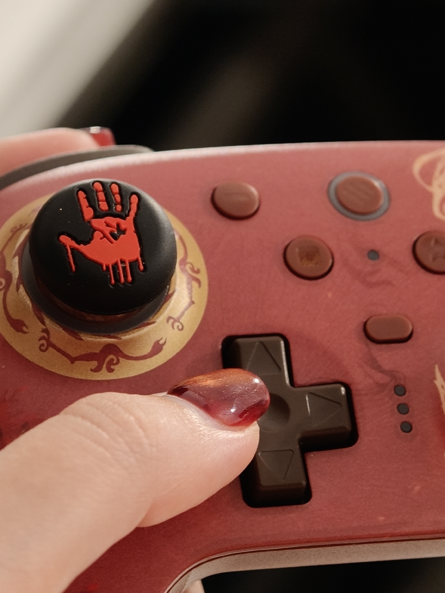 A close-up shot focuses on a person's thumb and index finger pressing down on a red video game controller. The controller features a black circular joystick with a red bloody handprint emblem. Decorative gold and red patterns adorn the controller's surface. The person's fingernails are painted with a glossy, reddish-brown polish, reflecting light. The background is dark and blurred, suggesting a dimly lit environment. There is no discernible information in the image that would indicate a location in Đà Nẵng, Vietnam.