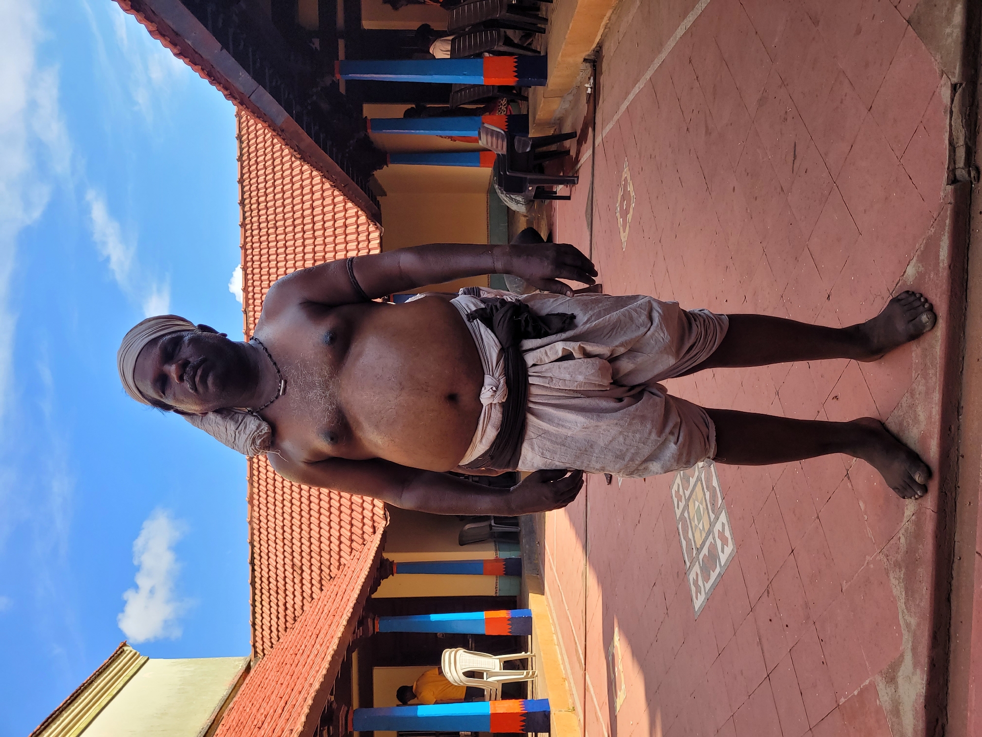 A bare-chested man stands centrally in a paved courtyard, looking directly at the viewer. He has a dark complexion, is of medium build, and wears a light-colored cloth wrapped around his head like a turban and another around his waist, resembling a dhoti. A dark beaded necklace is visible around his neck, and a dark band is on his left bicep. He stands barefoot on the reddish-brown tiled ground, which features some lighter diamond-shaped patterns.
The background reveals a traditional architectural setting under a bright blue sky with scattered white clouds. To the left and right, parts of a building with a distinctive reddish-orange tiled roof are visible, suggesting a covered walkway or veranda. Beyond the man, further into the building's interior, several structural pillars are brightly painted in blue and orange. Black and white plastic chairs are scattered in the shaded area. In one section, another person in a yellow shirt is partially visible, seated or crouched.
The bright lighting and clear sky suggest it is daytime. No specific activity beyond the man posing is clearly depicted. There is no visible text in the image. The architectural style hints at a location likely in South Asia, possibly a temple or a traditional home courtyard.