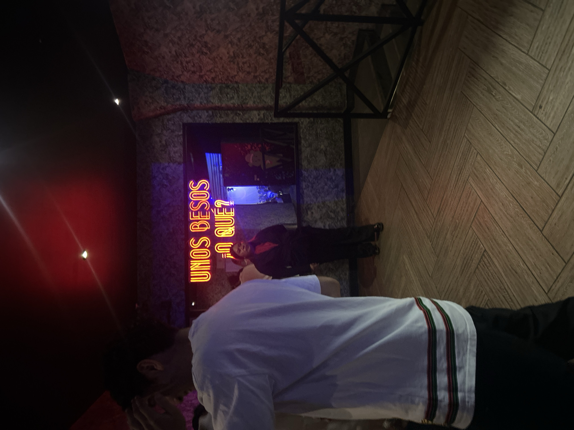 A slightly tilted indoor shot shows a man in a white shirt with green and red stripes on the sleeve, kneeling towards a neon sign that reads "UNOS BESOS ¿A QUE?" in red and purple colors. The shot is taken in what appears to be a bar or club-like environment. Behind the man, another person is visible standing near the neon sign, wearing a red shirt and a black blazer. A black framed picture is on the wall behind the standing person. The floor is herringbone wood parquet. A section of the ceiling shows a decorative metal frame. The left side of the image has a gradient of red, likely the ambience of the location. Based on the neon sign in Spanish, this photo was likely taken in Puebla, Mexico.