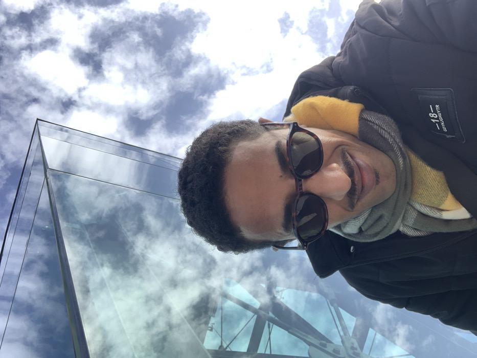 A low-angle outdoor shot frames a single individual, appearing to be a male, against a modern glass building and a partly cloudy sky. The individual, positioned centrally in the lower frame, has a medium-dark complexion, short dark curly hair, and a mustache. He wears dark, tortoiseshell-patterned oval sunglasses. His attire includes a black padded jacket with a white rectangular patch on the right side of the chest displaying text that includes "GDYKJGHUYTR -18". Around his neck is a multi-colored plaid scarf featuring prominent yellow, grey, and white segments. The individual's head is slightly tilted forward and downwards, displaying a subtle smile.
In the upper left background, a distinctive modern building constructed with reflective glass facades rises at sharp angles. The glass surfaces prominently reflect the blue sky and white cumulus clouds, effectively merging the building's form with the atmospheric conditions. This architectural style is consistent with the Tangier Med Port Center, located in Tangier, Morocco. The upper right background reveals an expanse of blue sky dotted with scattered white and grey clouds. The scene is brightly illuminated by natural daylight.