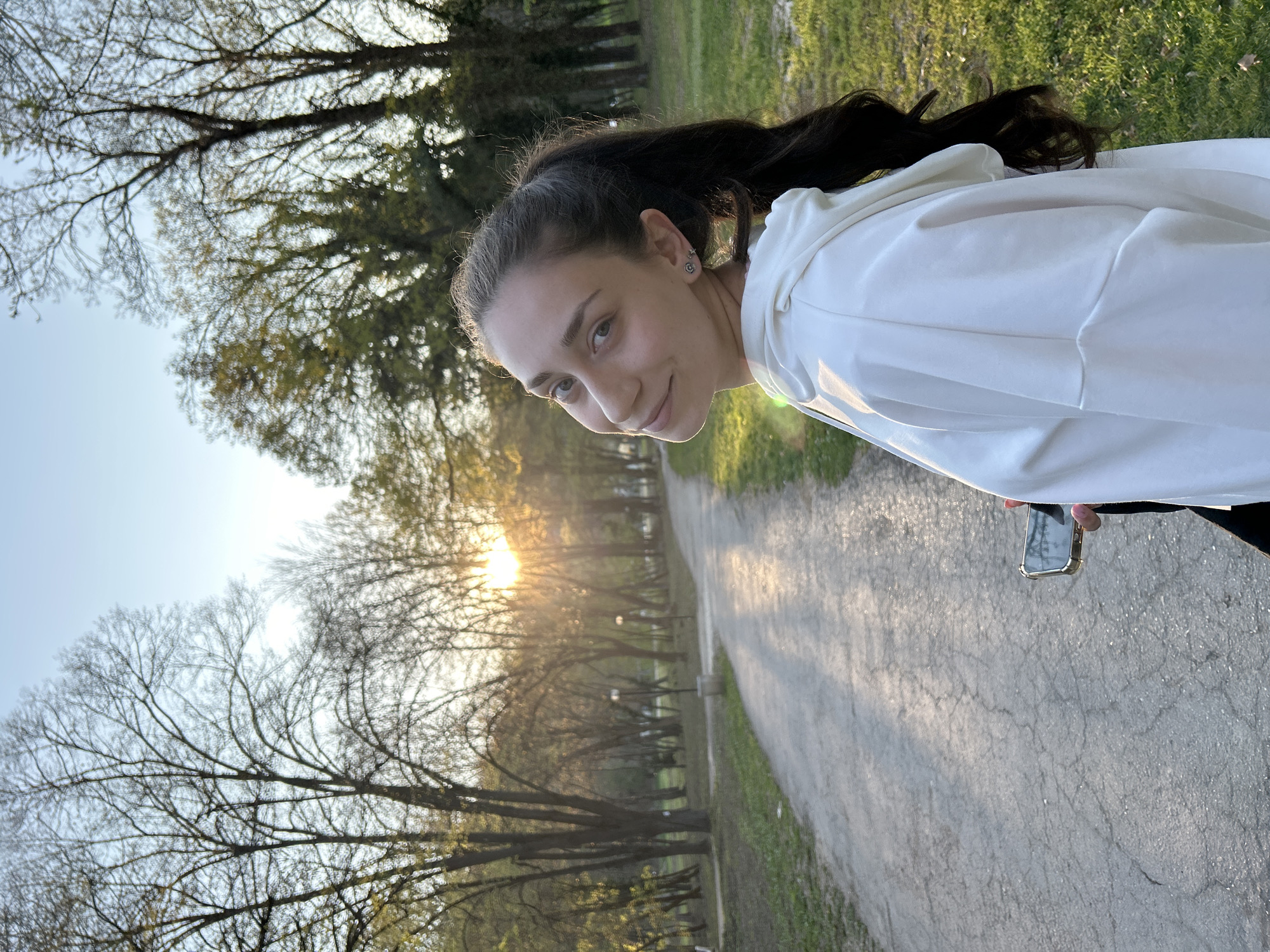 A young woman with brown hair pulled back into a ponytail and wearing a white hoodie is the primary subject of this photograph. She is facing the camera with a slight smile. She is standing on a paved pathway, holding a smartphone in her right hand, with a setting sun visible through the trees in the background. The pathway is lined with trees on either side, and there are patches of grass visible on both sides of the path. The location appears to be a park. Given the prompt's specific request for details, it's plausible this is a park located in Pleven, Bulgaria, based on the provided instruction. More context would be needed for definitive verification.
