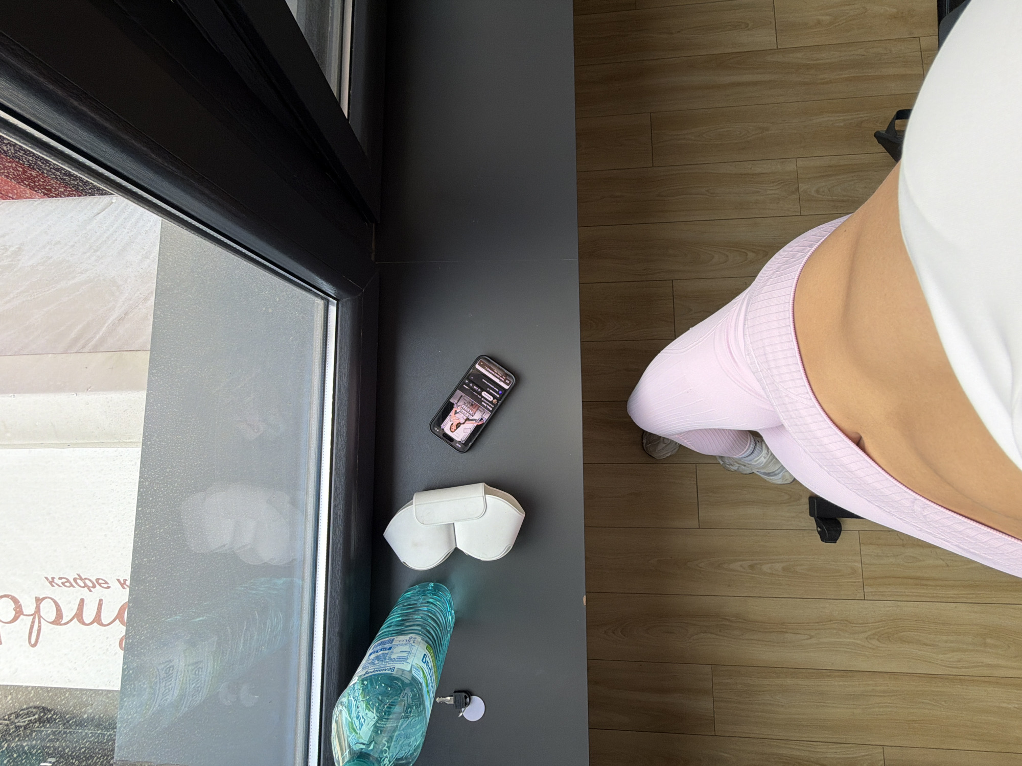 The scene is captured from a high-angle perspective, suggesting the camera is positioned above the objects and person in the frame. Part of a person wearing pink athletic leggings and a white top is visible on the right side of the image. The person is sitting on a black chair.
To the left, there's a window with some text visible on the glass, possibly indicating a cafe. The text includes the Cyrillic words "кафе к" followed by a word starting with "Ор". The window's external view is obscured.
Adjacent to the window and near the person, there's a dark gray surface. Resting on it are several items: a bottle of water with a blue label, a pair of white AirPods Max headphones, a black smartphone, and a key.
The floor appears to be made of light-colored wood panels.
The Cyrillic text "кафе к Ор" seen in the window suggests the location is likely in a country where the Cyrillic alphabet is used, such as Bulgaria. Further details on the water bottle label could confirm its origin. The city of Gabrovo, Bulgaria, is not determinable from this information.