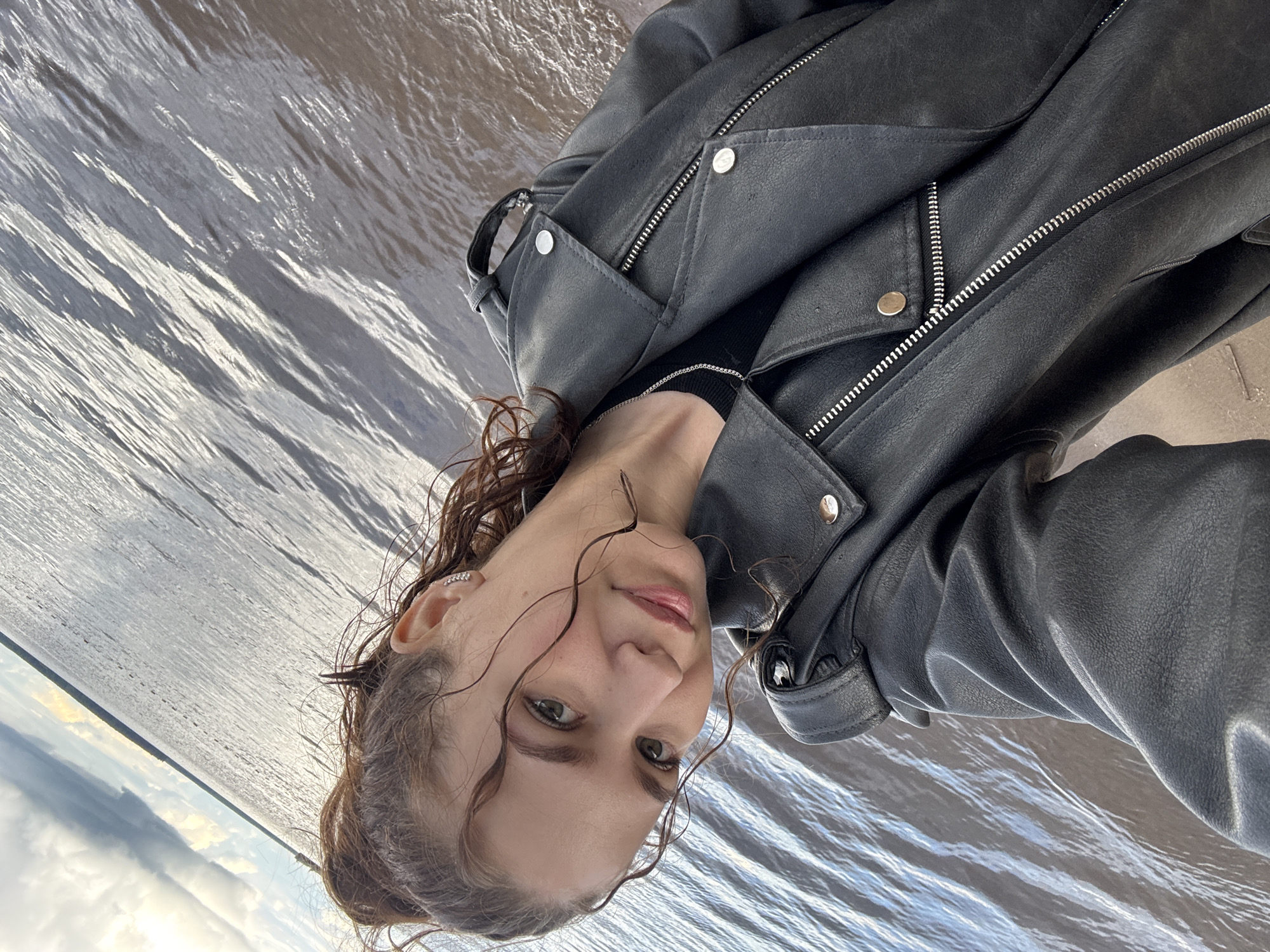 The image captures a close-up portrait of a young woman outdoors, looking directly at the camera. She has brown, wavy hair, with some wet strands clinging to her face, suggesting recent contact with water. Her light-colored eyes and subtly parted lips convey a calm, gentle expression. She is dressed in a black leather jacket featuring prominent silver zippers and snap buttons, worn over a black top, with a delicate silver chain necklace visible around her neck. A small, decorative ear cuff adorns her left ear.
The background is dominated by a large body of water, likely a river or lake, reflecting the sky and sunlight, creating a shimmering, rippled surface in shades of grey and silver. In the distance, a horizon line on the left separates the water from a partly cloudy sky, with lighter areas indicating the presence of sunlight. The image is composed on a diagonal axis, giving the scene a dynamic tilt.
The setting appears to be daytime, with soft, natural light illuminating the scene, possibly late afternoon. The woman's wet hair and the expansive body of water suggest she is at a natural outdoor location, possibly by the Kama River in Chaikovsky, Russia. There is no visible text in the media file.