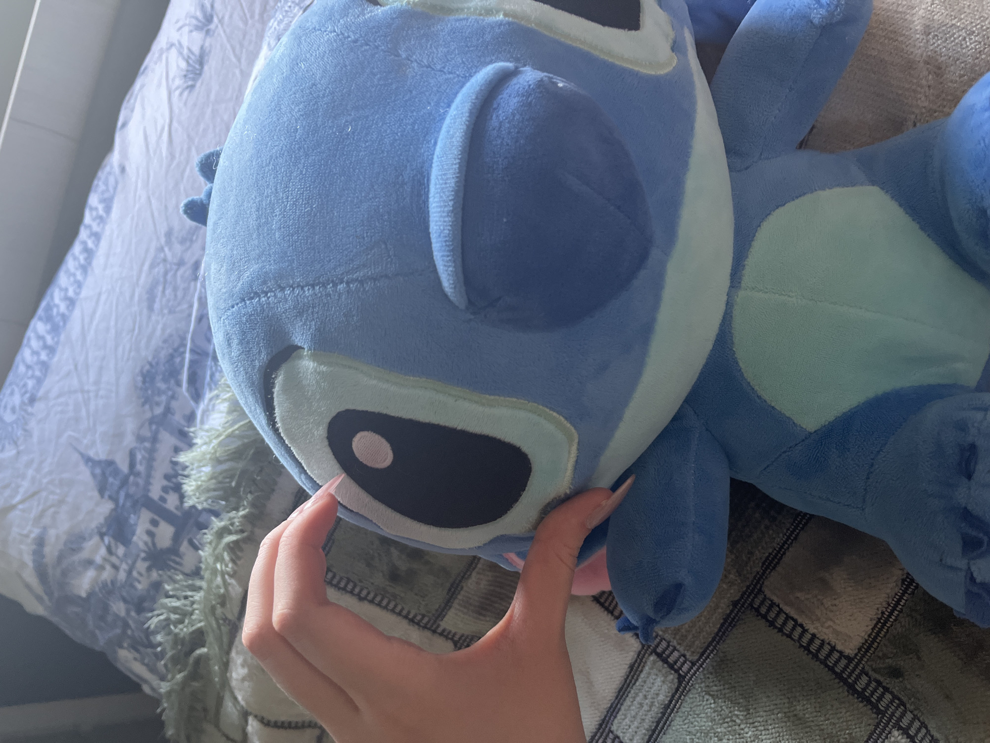 A close-up, high-angle shot shows a plush toy, identifiable as the character Stitch from Disney's Lilo & Stitch, resting on a bed. The toy is predominantly blue with lighter blue accents on its stomach and around its eyes. A human hand with manicured nails is visible on the left side of the frame, gently holding the toy's head. The background consists of a patterned bedspread with a blue and white design and some textured green fabric. The scene is indoors, with soft lighting. There is no indication of the city of Ashkadar, Russia.