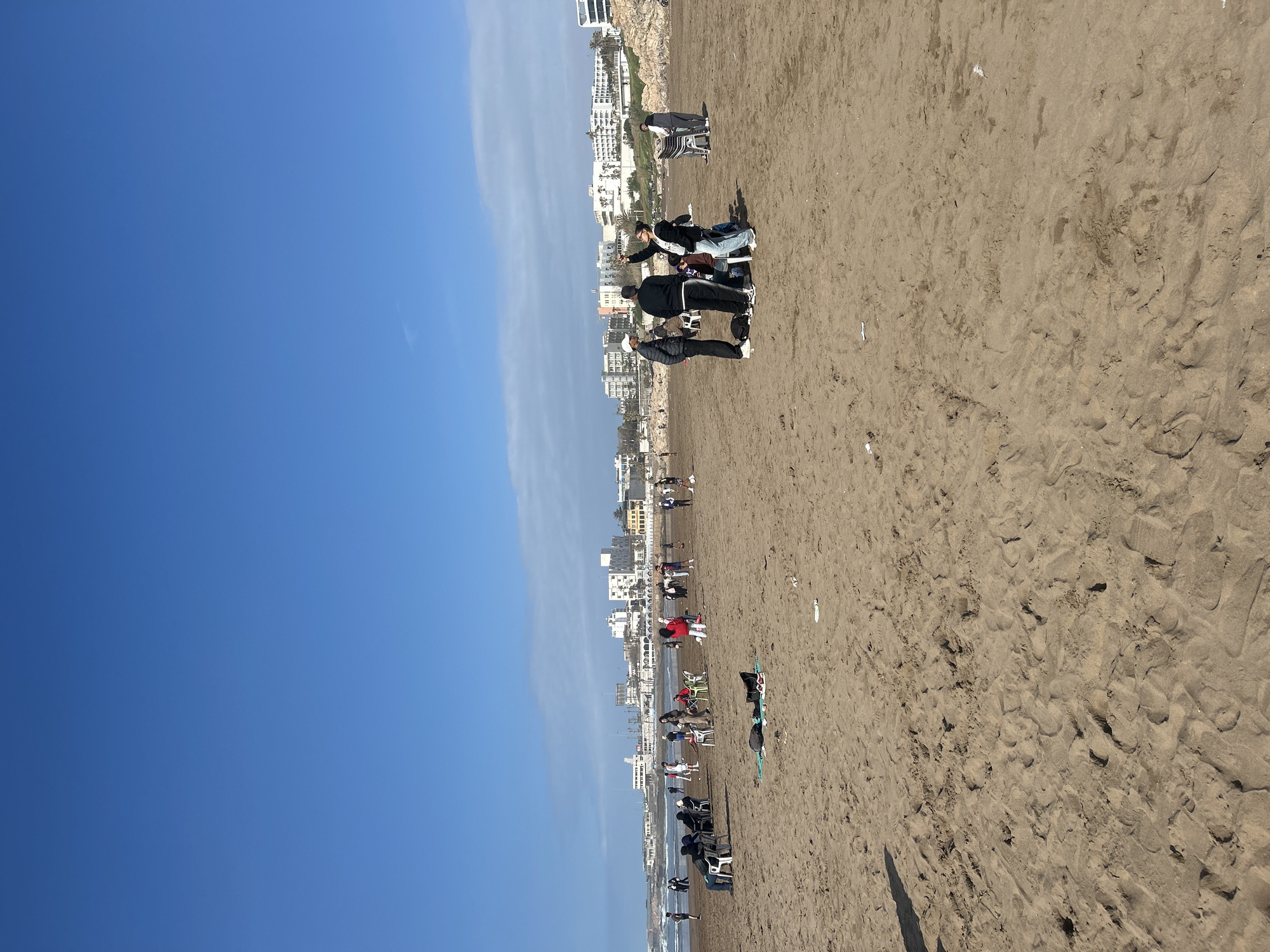 The image captures a sunny day at a beach in Casablanca, Morocco. The scene is oriented with the sky on the left and the sand on the right. A line of buildings runs parallel to the coastline, indicating urban development.
On the beach, groups of people are present. Some are standing in small clusters near the buildings, suggesting social interaction or photography. Other individuals or groups are further down the beach with chairs and personal items, indicating relaxation and leisure activities. The sand is a light brown color and shows footprints.
The sky is a clear blue, with a few wispy clouds near the horizon. The weather appears to be sunny and conducive to beach activities. Overall, the scene depicts a typical day at a popular beach location in Casablanca.