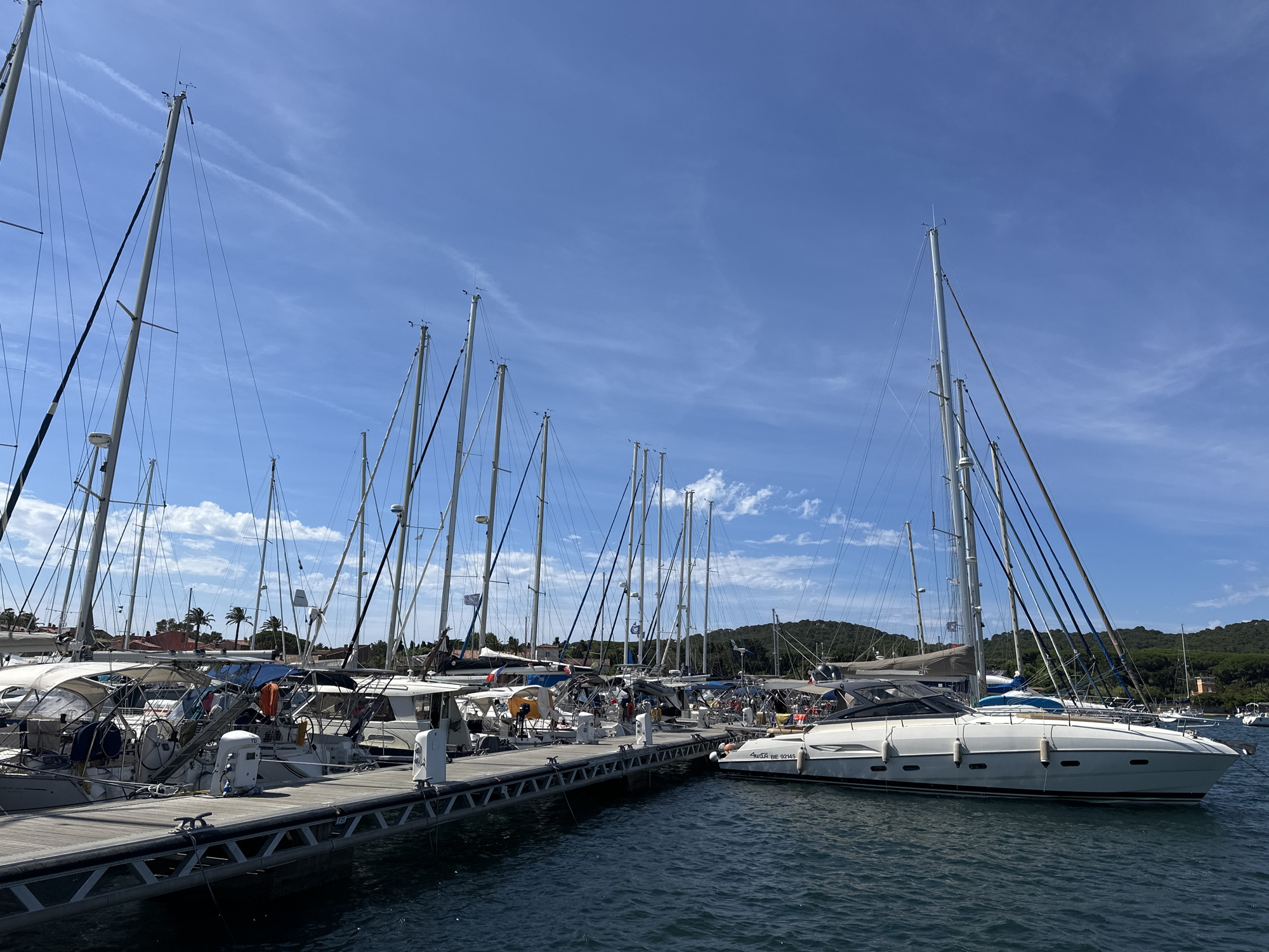 The image depicts a sunny day in the port of Hyères, France, showcasing a marina filled with numerous sailboats docked along a wooden pier. The sky is a clear blue with a few wispy white clouds. The sailboats, varying in size and design, are moored side-by-side, with their tall masts and rigging dominating the upper portion of the frame. The water is calm and reflects the blue of the sky. On the pier, various nautical equipment such as cleats, ropes, and fenders are visible. In the background, lush green hills with scattered buildings and palm trees suggest the Mediterranean coastline. A few modern motorboats are also present amongst the sailboats. The overall scene conveys a sense of a peaceful and active marina on a pleasant day.