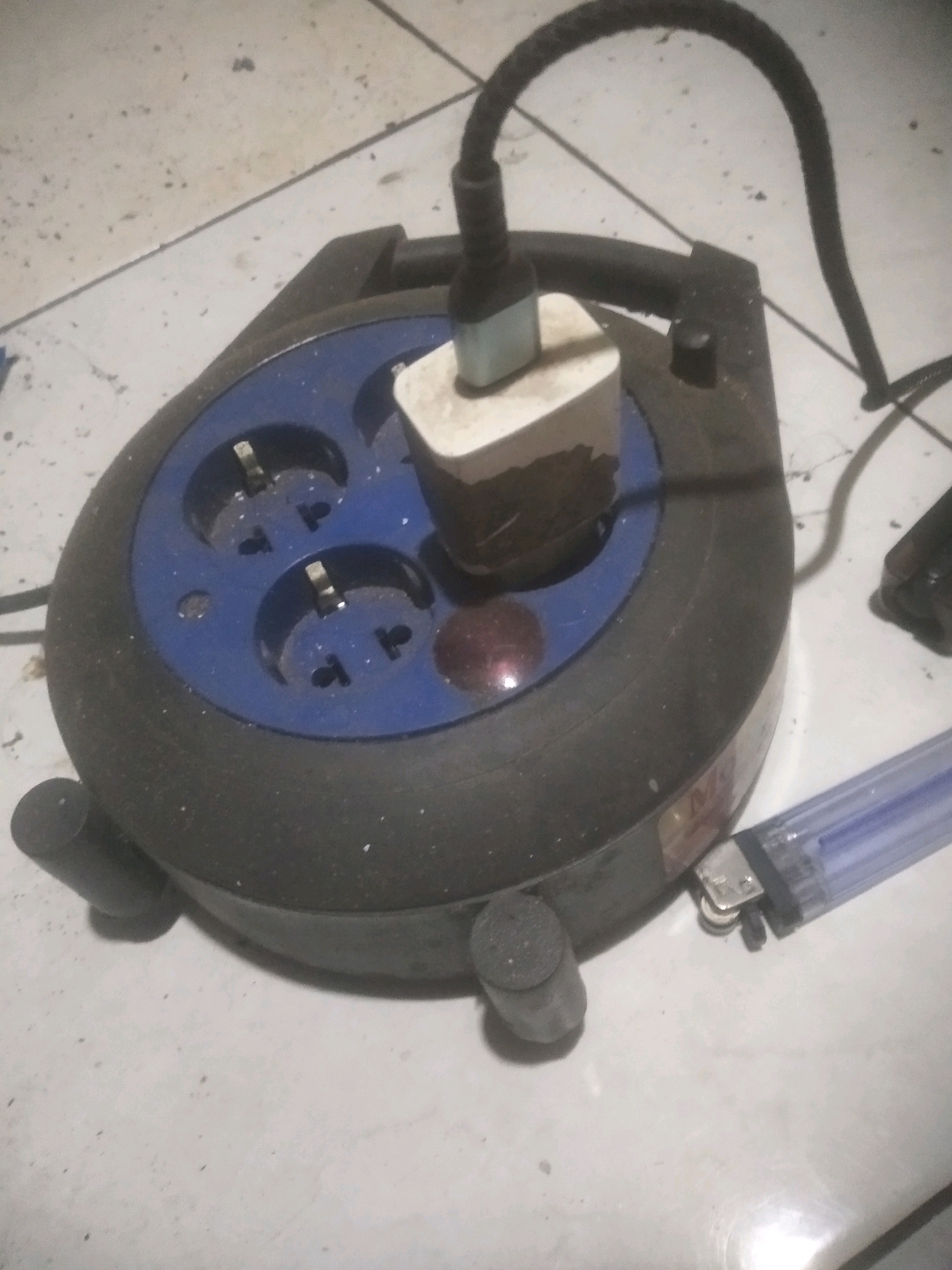 The image depicts a dark, circular extension cord reel with multiple universal electrical outlets, one of which is occupied by a white and blue electrical plug. The reel is situated on a light-colored, tiled floor, which appears somewhat dirty with scattered dark specks. To the right of the extension cord reel, a clear plastic lighter with a blue flame chamber is visible. The overall lighting is dim, casting shadows and highlighting dust on the surfaces. The scene appears to be an indoor setting, possibly a room or a workshop, and is consistent with common household environments found in Cilegon, Indonesia.