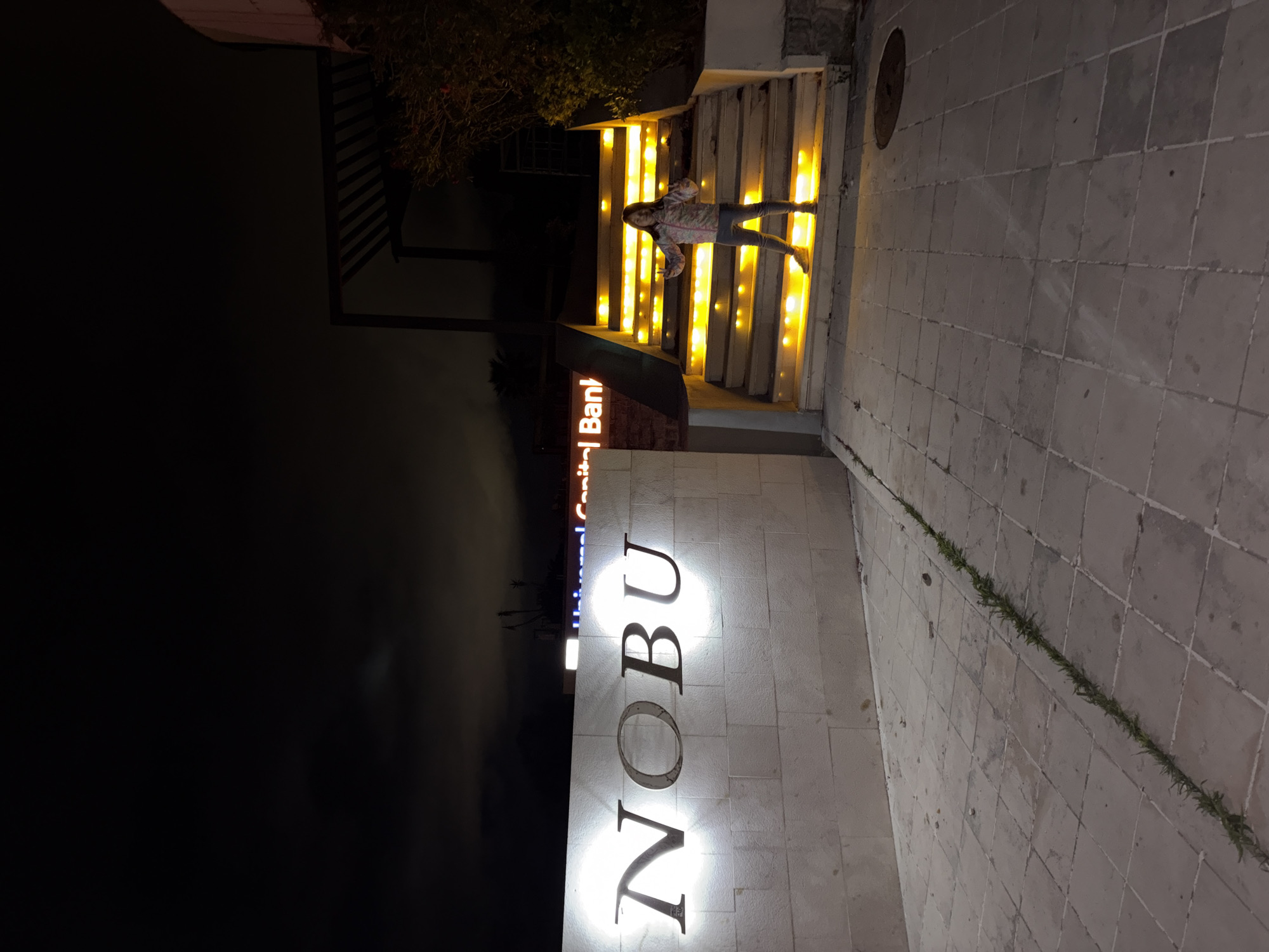 A dimly lit, rotated, medium-shot photograph captures the exterior of the Nobu restaurant in Sveti Stefan, Montenegro, at night. A young person is lying on a staircase illuminated by yellow lights. The person is wearing a patterned hooded jacket and pants. The name "NOBU" is spelled vertically in illuminated block letters on a light-colored wall to the left of the staircase. Above "NOBU", a sign with the words "Central Capital Bank" is partially visible. A stone-paved ground stretches out in front of the building.