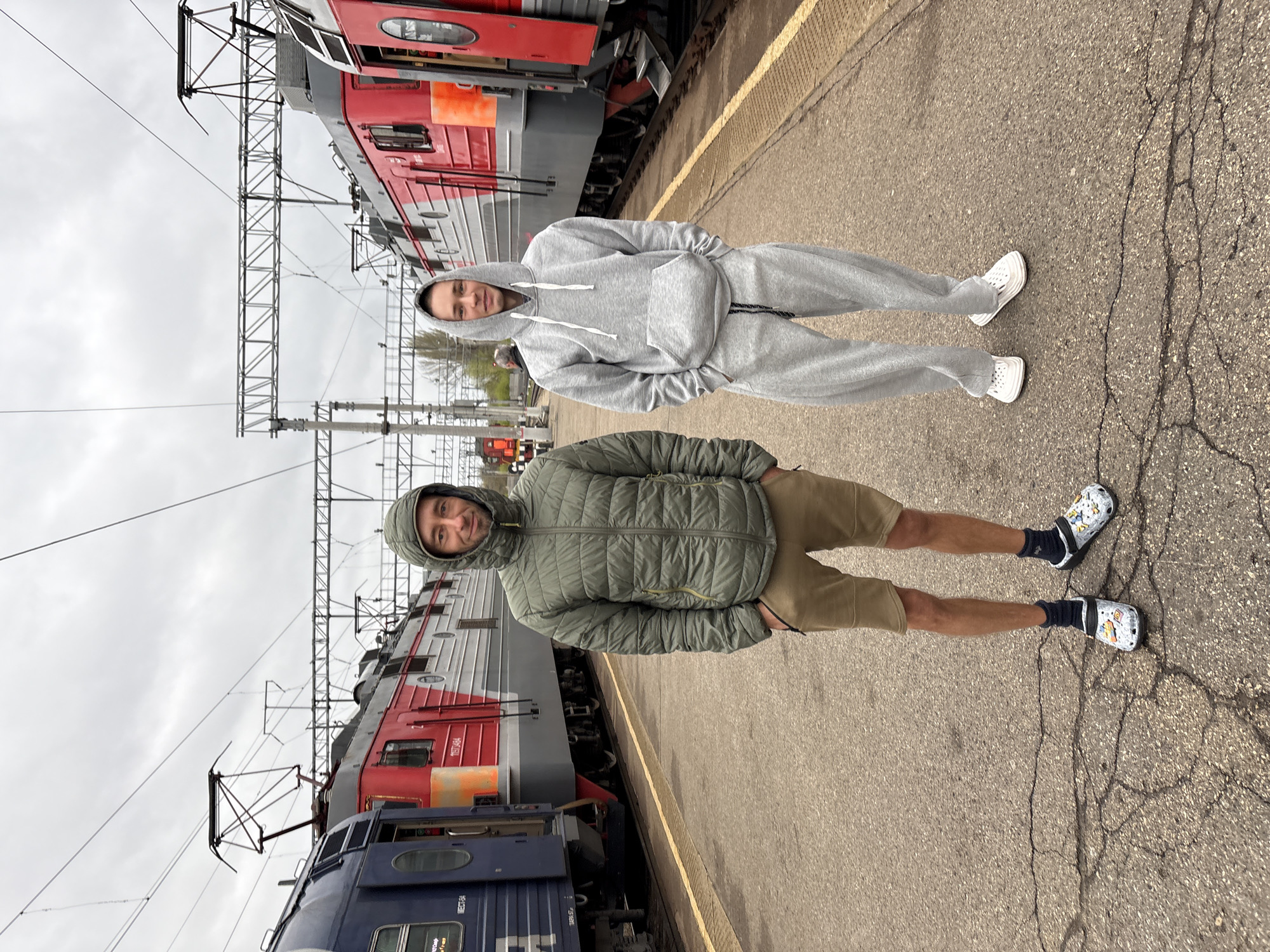 Two men stand on a railway platform in Saratov, Russia. The man in the foreground wears a green quilted jacket, khaki shorts, black socks, and white patterned Crocs. The man in the background is wearing a grey tracksuit and white sneakers. Behind them are several train locomotives with red, grey, and blue color schemes. Overhead, electrical cables and supporting structures run along the tracks. The platform is cracked and uneven, and the sky is overcast.