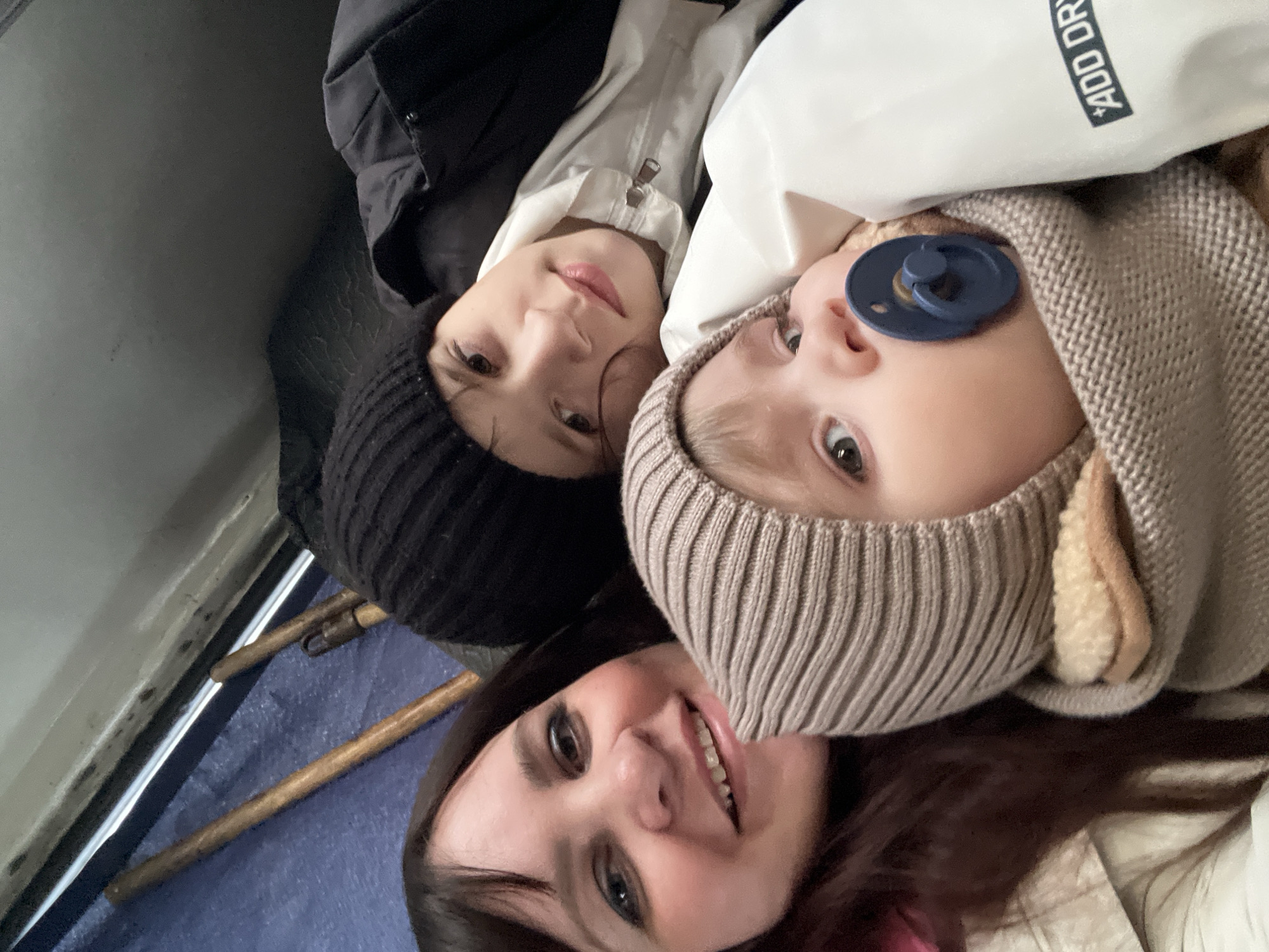 Three individuals are positioned close together, likely for a photograph. The person at the bottom of the frame is a female with dark hair and a smiling expression. Above and to the left is a child wearing a black knit hat and a dark jacket. A baby is positioned above and to the right. The baby is wearing a knitted hat and has a pacifier in their mouth. They are indoors, possibly in a vehicle. There is no evidence in the image to confirm they are in Noviy, Russia.
