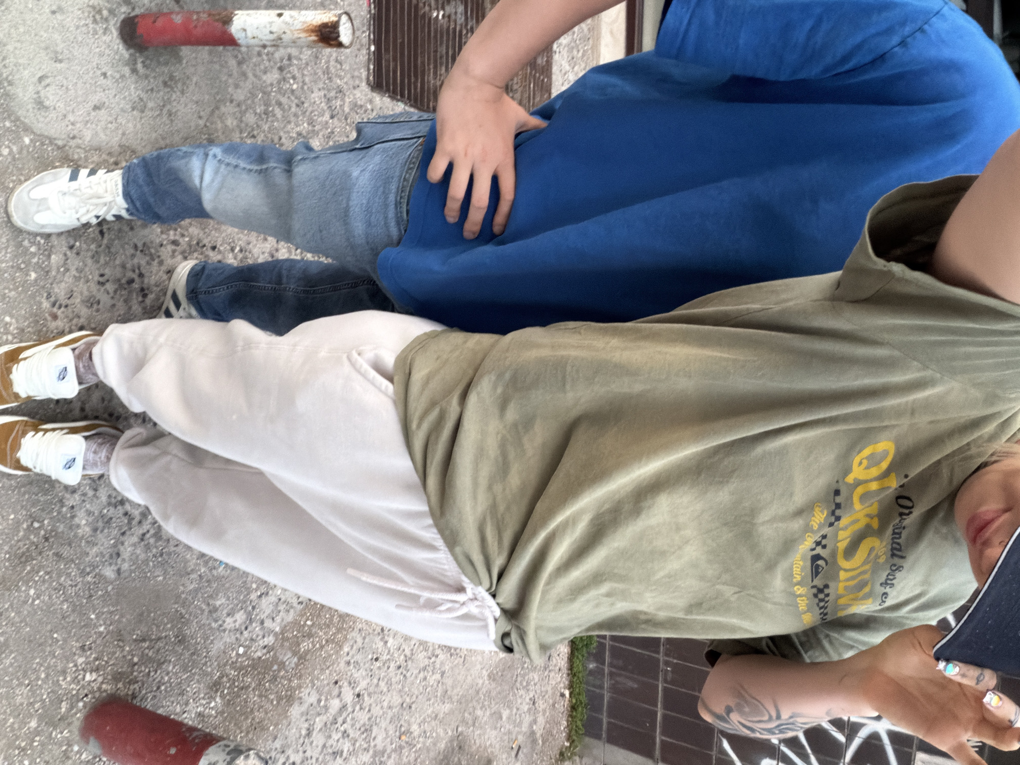 The image depicts two individuals standing on a cracked concrete surface, possibly a sidewalk or pedestrian area. To the left, there is a short, damaged metal post painted red and white. One person wears light-wash denim jeans and white sneakers with blue stripes, with a blue hooded sweatshirt. Their hand rests on top of the second individual, who wears beige sweatpants and brown sneakers. The second person has a green t-shirt, and their forearm reveals a tattoo. Part of their face is visible, and they are holding a mobile device. The setting appears to be in an urban environment, and based on the photo's origin, it's plausible the location is Budva, Montenegro. The exact interaction between the people is unclear but they are standing near each other.