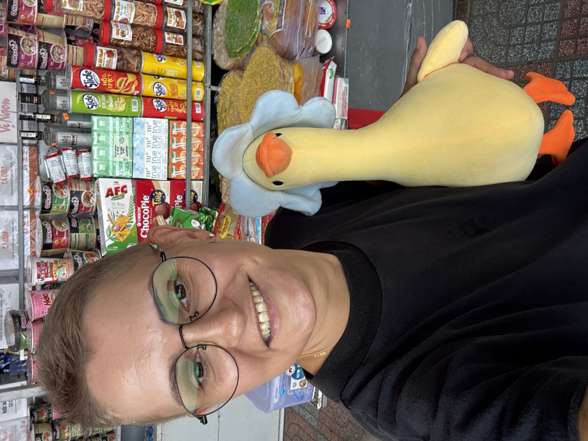 In an outdoor market stall in Nha Trang, Vietnam, a person with short, brown hair and glasses is taking a selfie, holding a yellow duck plush toy with a blue flower hat. The person is wearing a black t-shirt. The background is filled with shelves stocked with various food items, including instant noodles, snacks, and bottled drinks. The stall appears to be well-lit and bustling with merchandise.