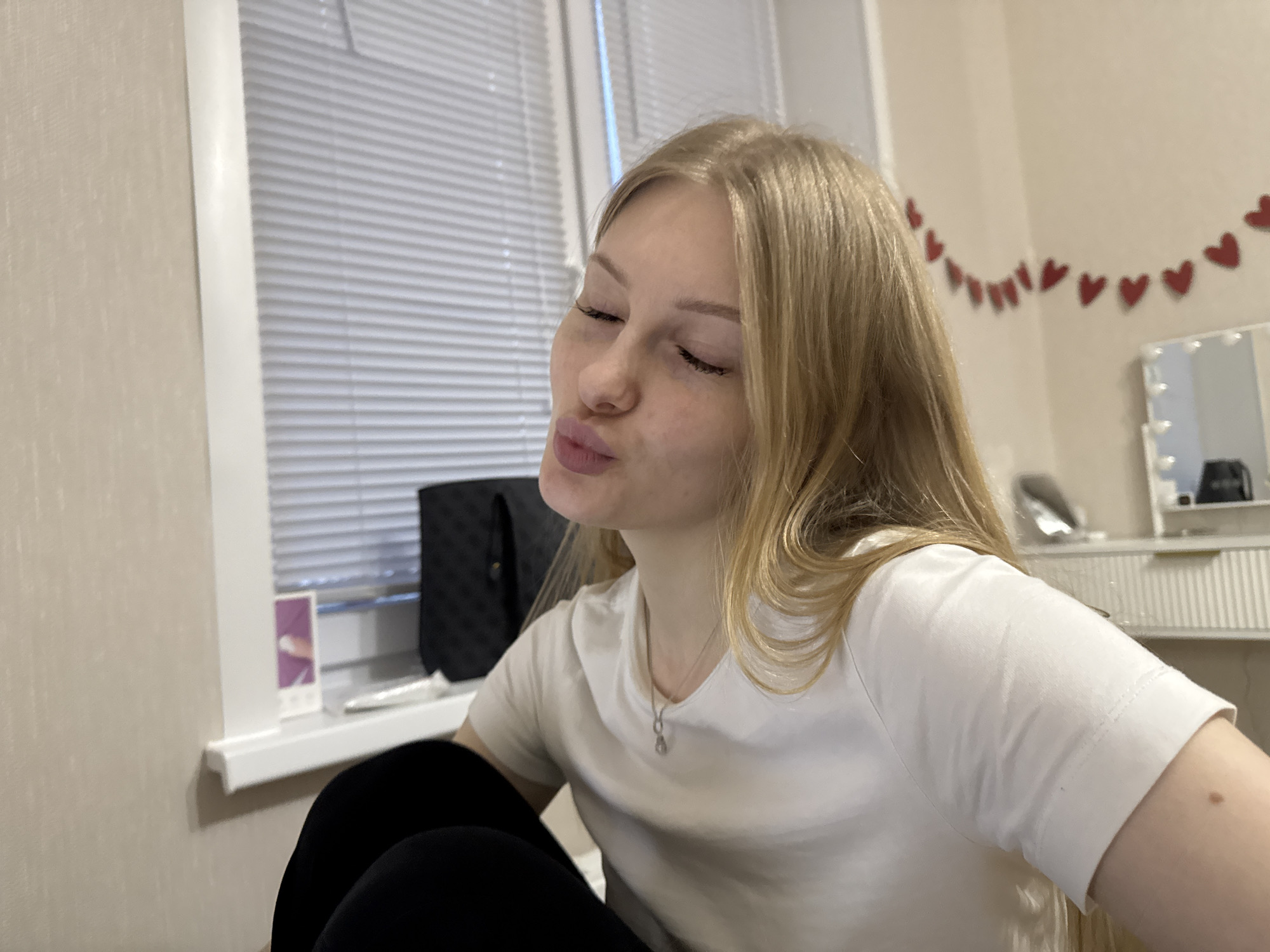 A young woman with long, straight blonde hair and fair skin is depicted in a medium-close-up, indoor shot. She is positioned facing slightly to the left, with her eyes closed and her lips puckered in a playful or "kissing" expression. She is wearing a plain white short-sleeved t-shirt and dark-colored leggings. A delicate silver necklace with a small, elongated pendant is visible around her neck.
The setting is an indoor room, likely a personal space such as a bedroom, located in Perm, Russia. To the left of the woman, a window is visible with light-colored horizontal blinds, allowing diffused light to enter the room. A white windowsill runs beneath the window, on which a black textured bag and a small, upright rectangular object (possibly a card or photo frame) with a purple/pink design can be seen. The wall to the left of the window and behind the woman is covered in light beige, textured wallpaper.
In the background to the right, a string of red heart-shaped decorations is hung along the wall. Further right, a white vanity table with an illuminated mirror, featuring visible light bulbs around its frame, is partially visible. The overall lighting is soft, suggesting indoor artificial light or subdued daylight filtering through the blinds. The woman's pose and expression convey a lighthearted and affectionate mood. No legible text is discernible in the image.