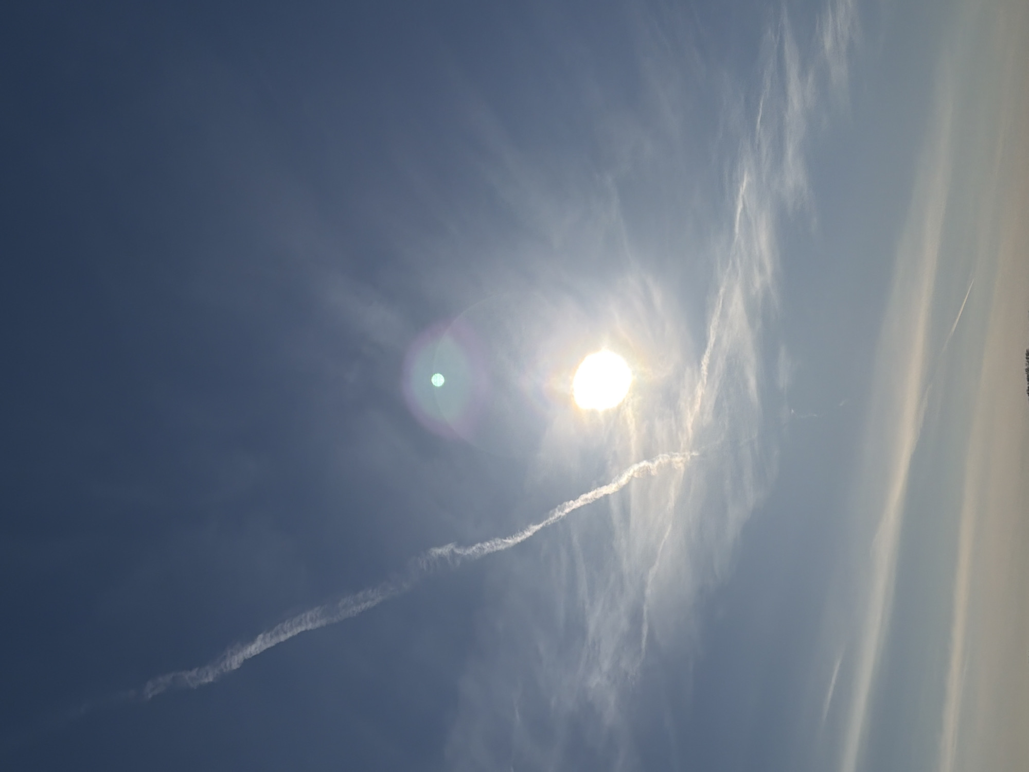 The image shows a daytime view of the sky. A bright sun is visible with lens flare. There are scattered clouds, including a contrail of an aircraft cutting across the sky from lower left towards the sun. The sky is blue. The photograph appears to have been taken outdoors. There is no information in the image indicating that the photograph was taken in Votkinsk, Russia.