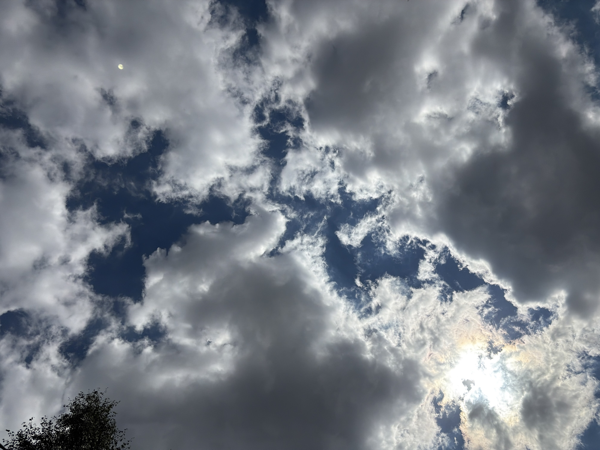 This is a photograph of a cloudy sky taken from the city of Imeni Vorovskogo, Russia. The image shows a dynamic sky with a mix of dark and light clouds, with patches of blue sky visible. The sun is partially obscured by clouds, creating bright highlights and some lens flare. A faint disc, possibly the moon, is visible in the upper right quadrant of the image. A silhouette of tree branches is visible in the upper left corner. The perspective is from below, looking upwards. There are no people or man-made structures clearly identifiable in the scene, other than the implied location of the photographer within the city.