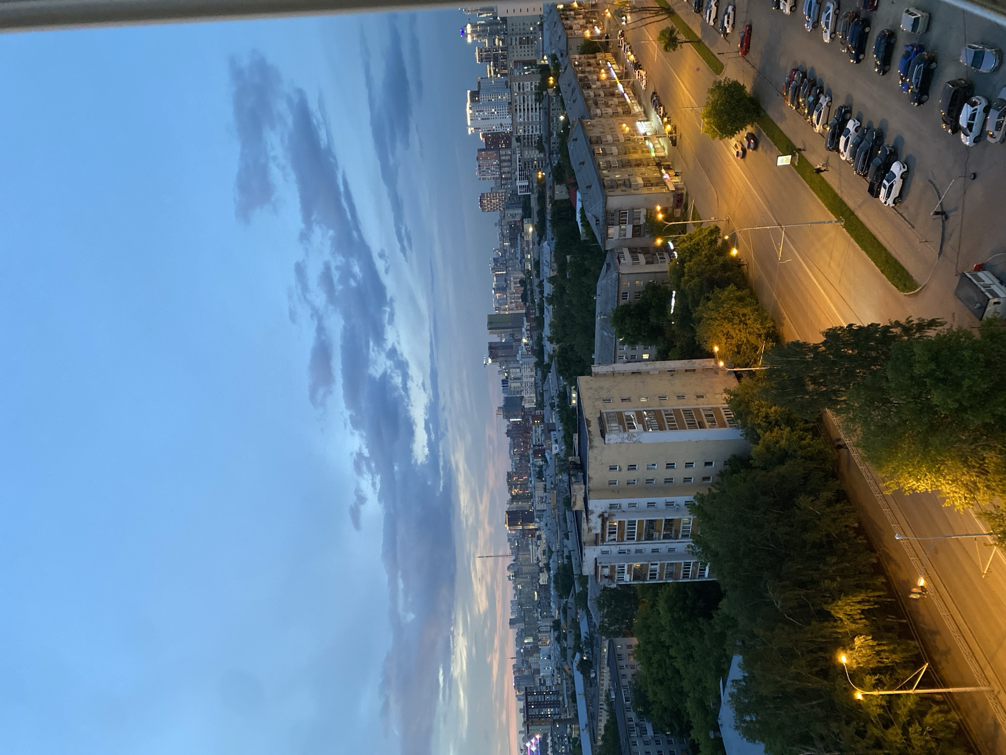 The image, taken from an elevated perspective, captures an urban landscape in Yekaterinburg, Russia, during dusk or early evening. The scene is oriented vertically, with the sky on the left and the city extending across the right.
The sky is a mix of clear blue and scattered, elongated clouds, some appearing wispy while others are more substantial. Faint hints of pink and orange are visible along the horizon line where the sky meets the distant cityscape, suggesting the last vestiges of sunset.
The urban environment on the right is extensively lit by artificial sources. A wide, multi-lane road stretches through the mid-ground, featuring numerous streetlights that emit a warm, yellow glow, illuminating the asphalt and surrounding areas. Along the right side of this main road, a significant number of cars are parked in rows, including various models and colors such as white, black, blue, and silver sedans and SUVs. A smaller road is visible further left with additional parked vehicles. At the far right edge, a white utility or delivery truck is parked.
A dense array of multi-story buildings populates the cityscape, ranging from older structures with visible balconies and varied facades to more modern, taller buildings in the distant background. Many windows in these buildings are lit from within, suggesting occupancy. Green trees are interspersed throughout the urban fabric, lining the roads and providing pockets of foliage amidst the concrete.
No explicit human activity is clearly visible, but the illuminated buildings and numerous parked vehicles indicate an active urban setting settling into the evening. The overall atmosphere is one of a calm, well-lit city transitioning from day to night. The top of the image is partially obscured by a light-colored window frame or balcony railing. No legible text is discernible within the image.