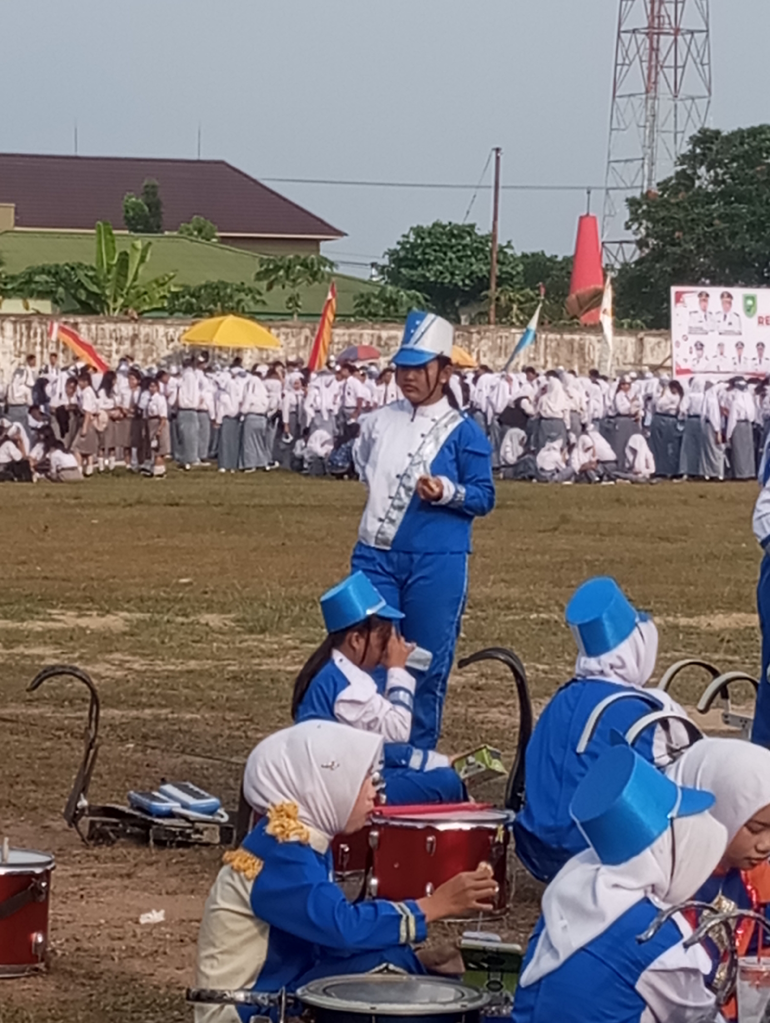 In Candirejo, Indonesia, a group of young people in marching band uniforms are gathered outdoors on a grassy field. The central figure, wearing a blue and white uniform with a blue cap, stands and appears to be addressing or leading the group. Several other individuals, also in matching blue and white uniforms, are positioned around her, some seated and others standing. Some are wearing white headscarves. Musical instruments, including drums, are visible on the ground and being held. In the background, a crowd of people in school uniforms are visible, along with buildings, trees, and a large radio tower. The sky is overcast.