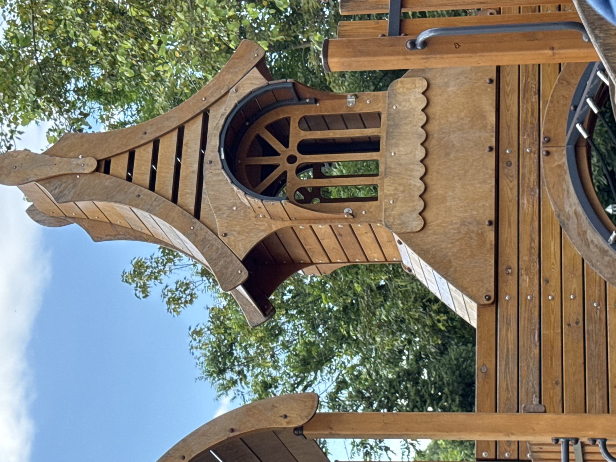 The image shows a close-up of a wooden playground structure in Верхнерусское, Russia. The structure features an ornate wooden tower with a peaked roof and a unique arched window with decorative wooden slats. The tower is built with horizontally placed wooden planks and is supported by vertical wooden posts. In the background, lush green trees are visible against a bright blue sky with scattered white clouds. Part of another wooden playground element is visible on the left side of the frame. A black metal handle and a small heart shape are visible on the right side of the structure. The scene is captured in daylight, suggesting an outdoor setting, likely a park or a dedicated playground area.