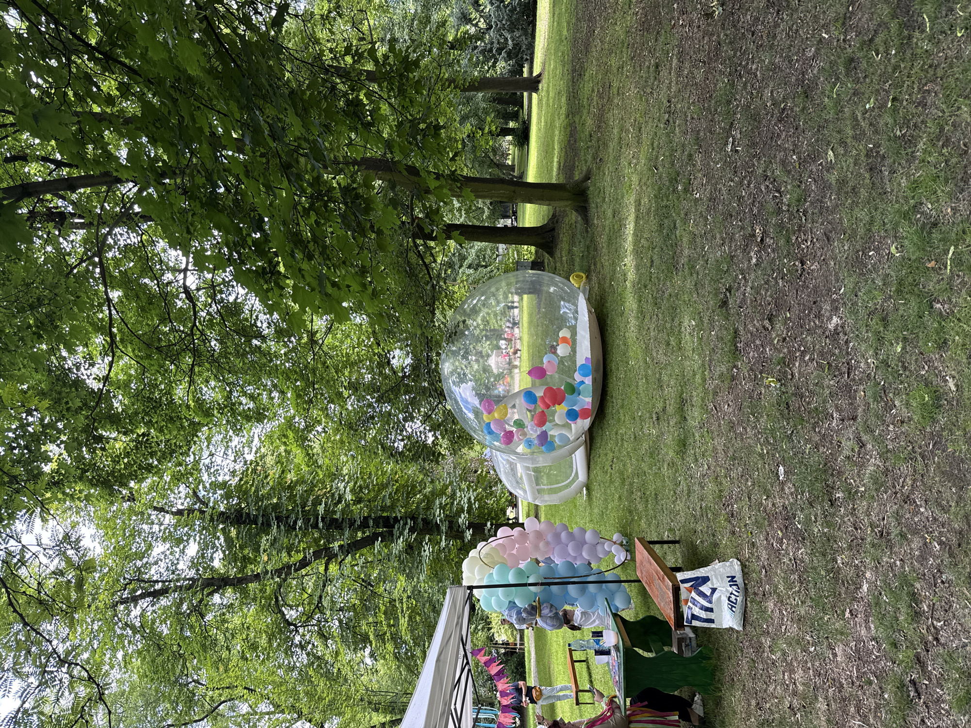 The image depicts an outdoor event taking place in a park-like setting during daylight hours. The location is characterized by a dense canopy of mature trees with vibrant green foliage and a wide expanse of green grass, some areas of which show signs of wear and scattered debris.
In the left foreground, a white canopy tent is erected. Beneath the tent, a green-painted wooden picnic table and a wooden bench are positioned. On the table, a clear plastic water bottle and a white cup are visible, alongside some papers or magazines. Partially seated at the table, with their back to the camera, is a person whose arm is adorned with colorful ribbons. Another individual is partially visible standing behind a large, pastel-colored balloon arch. This arch, forming a circular or oval shape, is composed of numerous balloons in shades of light blue, mint green, white, pink, and lavender. A white shopping bag with the blue text "TIN ACT.N" (likely "ACTION") rests on the ground underneath the wooden bench. Further behind the tent, a person wearing a black top and light-colored pants with a yellow fanny pack is standing.
In the midground, two clear inflatable structures are situated on the grass. The larger, dome-shaped structure on the right is filled with a multitude of brightly colored balloons, including red, blue, yellow, green, pink, purple, white, and orange, suggesting a play area. A smaller, empty inflatable dome is connected to the larger one by a transparent tunnel. Beyond these structures, several tree trunks are visible, indicating more trees further into the park.
The background shows additional expanses of green grass and numerous trees, their canopies creating a lush, verdant environment. Faint figures of people are discernible in the far distance through the clear inflatable bubble, implying more activity further away. The overall lighting suggests a sunny or partly cloudy day.