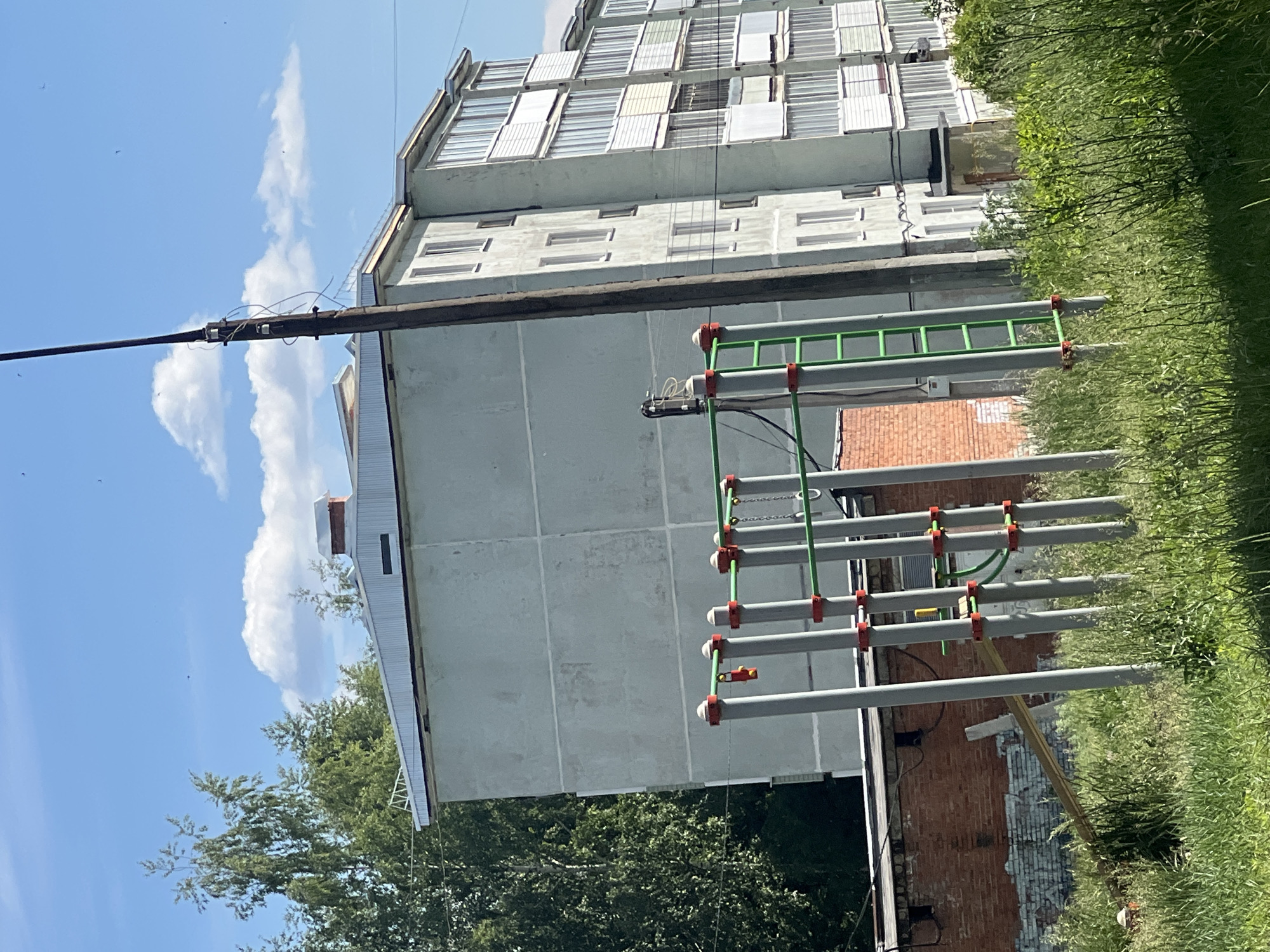 This forensic description details an outdoor scene in the city of Noviy, Russia, depicting a residential area under a clear blue sky with scattered cumulus clouds.
In the immediate foreground, a patch of unkempt, tall green grass obscures the base of various structures. An outdoor children's gym or workout area occupies the central foreground, consisting of multiple grey vertical metal poles topped with red caps and featuring red bands connecting to green horizontal bars and ladders. A red and yellow object, possibly a toy or part of the equipment, hangs from one of these poles. To the left, partially hidden by the grass, lies a light-colored wooden plank or beam leaning against a structure, and a small, partially obscured orange/brown object is visible on the ground.
In the midground, a single-story, dilapidated red brick building with dark rectangular openings (possibly windows or vents) and areas of peeling white or light blue paint/graffiti is visible on the left. Dominating the right half of the image is a multi-story (at least five visible floors), light grey/pale green apartment building constructed with concrete panels, showing visible seams and weathering. The building features numerous white-framed windows and multiple enclosed balconies, predominantly white, projecting from its facade. A tall, dark brown utility pole with attached wires, some appearing loose, stands prominently in front of the apartment building, slightly left of center. Behind the main apartment building, the light-colored corrugated metal roof of another structure is visible, topped with a small red brick chimney.
The background is filled with dense green deciduous trees and some conifers, particularly on the left. The sky is bright blue, with several small, dark specks, possibly birds or insects, visible against it. No people are present in the scene. The overall impression is an older, somewhat weathered residential area.