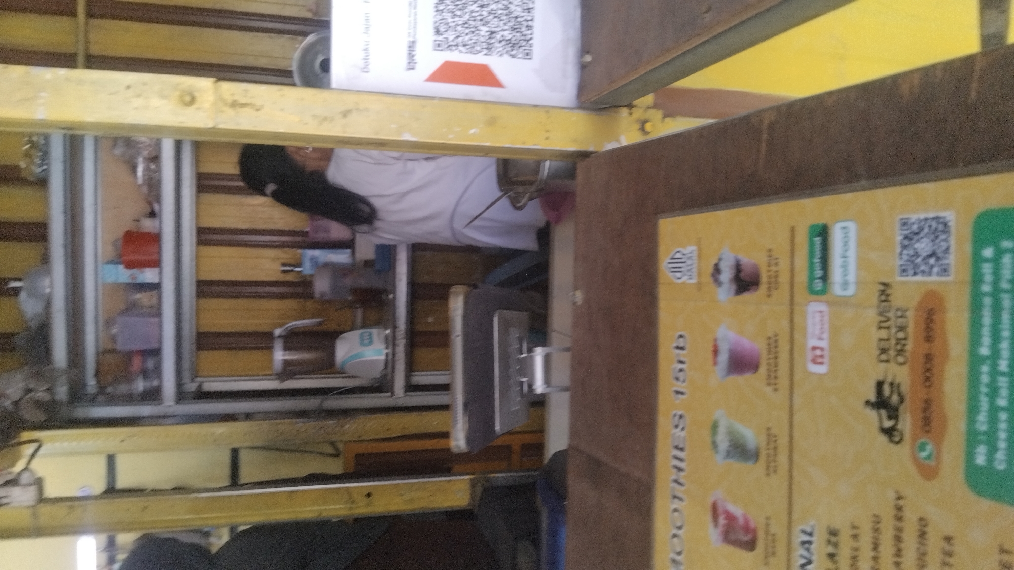 This is an eye-level, medium shot of a food stall in Proyonangg, Indonesia. The stall appears to be selling smoothies and other food items. A young woman with dark hair, wearing a white shirt and black pants, is working behind the counter, preparing a drink. The counter is made of wood and has a yellow menu board with pictures of smoothies and prices. There are also logos for food delivery services like GoFood and GrabFood. The background shows shelves with various products, including drinks and containers. A QR code is visible on a sign near the counter, likely for payment or ordering. The overall scene depicts a typical Indonesian food stall, bustling with activity during a daytime operation.