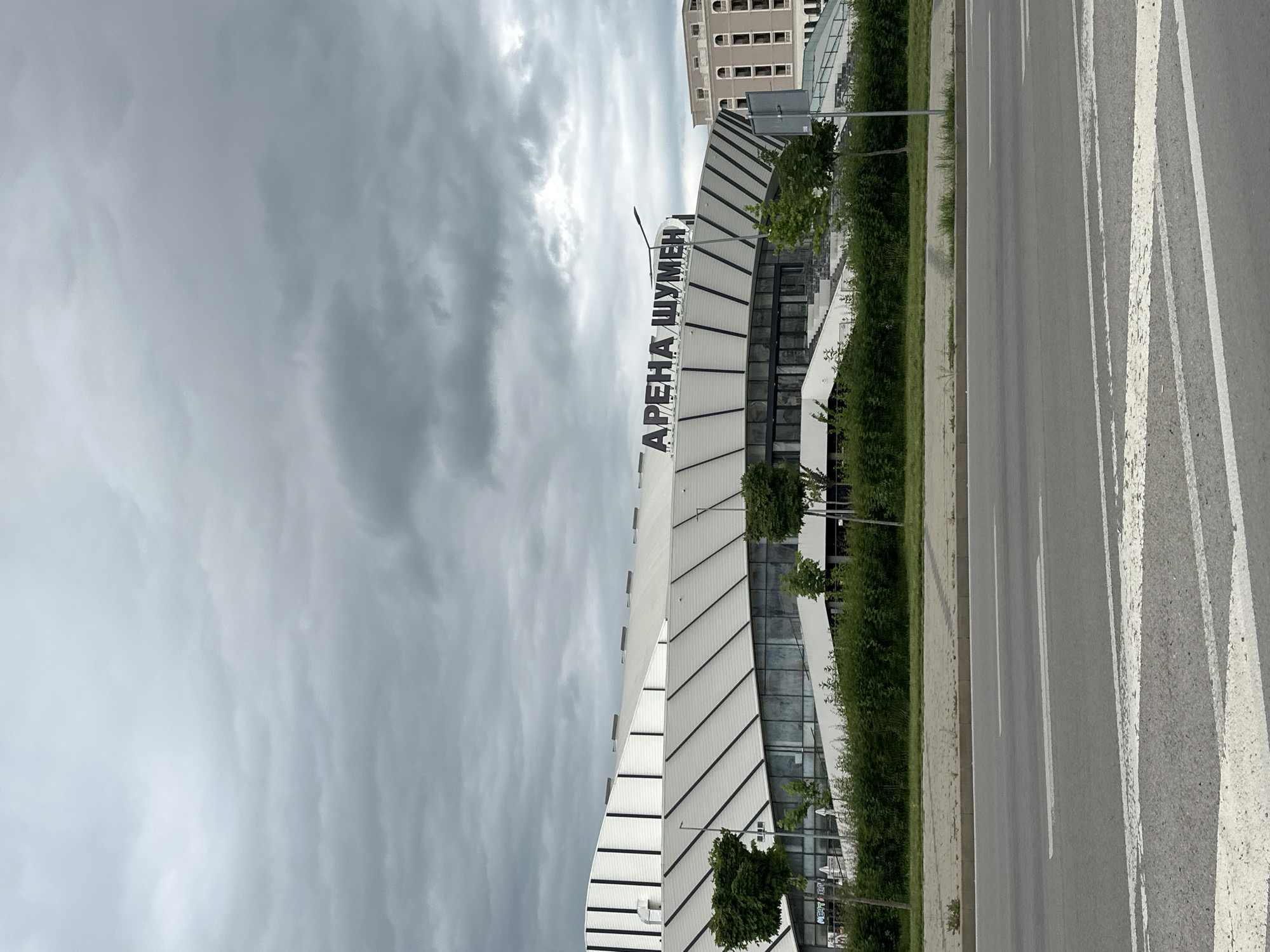 The image shows Arena Shumen, a sports and entertainment complex in the Bulgarian city of Shumen. The modern building has a curved, mostly white facade with black accents, and the words "Arena Shumen" are vertically displayed on the facade. The Arena is situated along a road marked with white lines for traffic. A green median with small trees separates the Arena from the road. In the background, a multi-story building can be seen. The sky above is overcast with gray clouds, indicating cloudy weather. There are no people visible in the image.