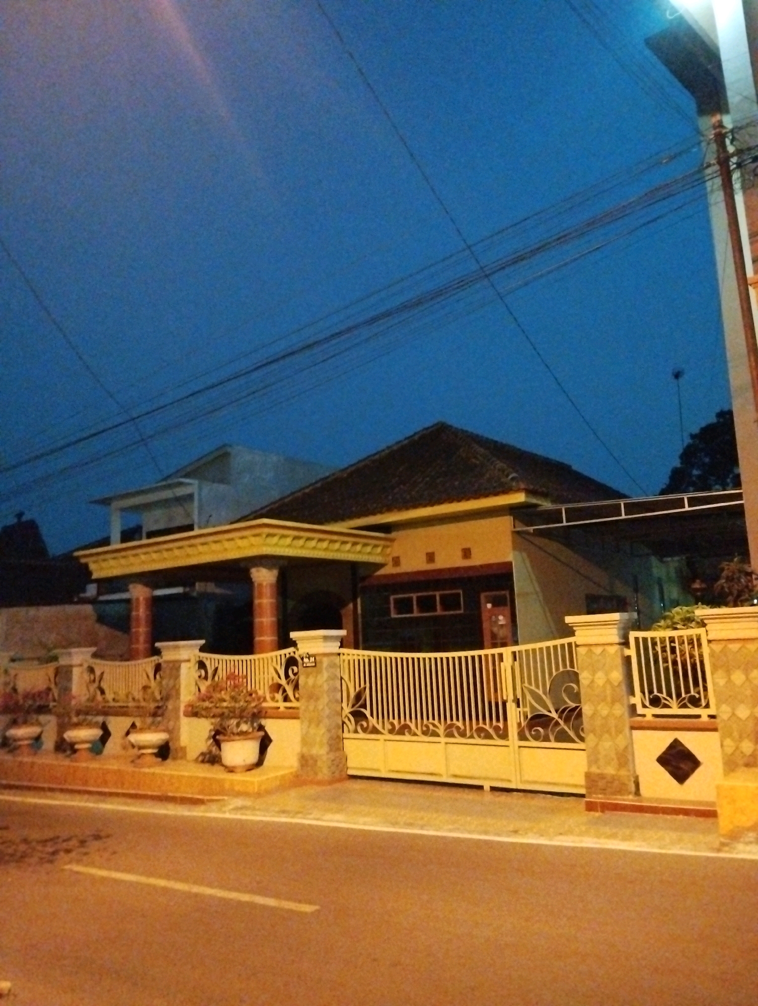 The image shows a house with a fence and a road in front of it, at dusk. The house is a single-story building with a brown tiled roof and yellow walls. It has a covered patio with pillars in the front. The fence is made of metal with decorative elements. There are potted plants in front of the fence. The road is paved and has a white line marking the lane. The sky is dark blue, indicating it is either early morning or late evening. The overall scene is that of a residential area in Salatiga, Indonesia.