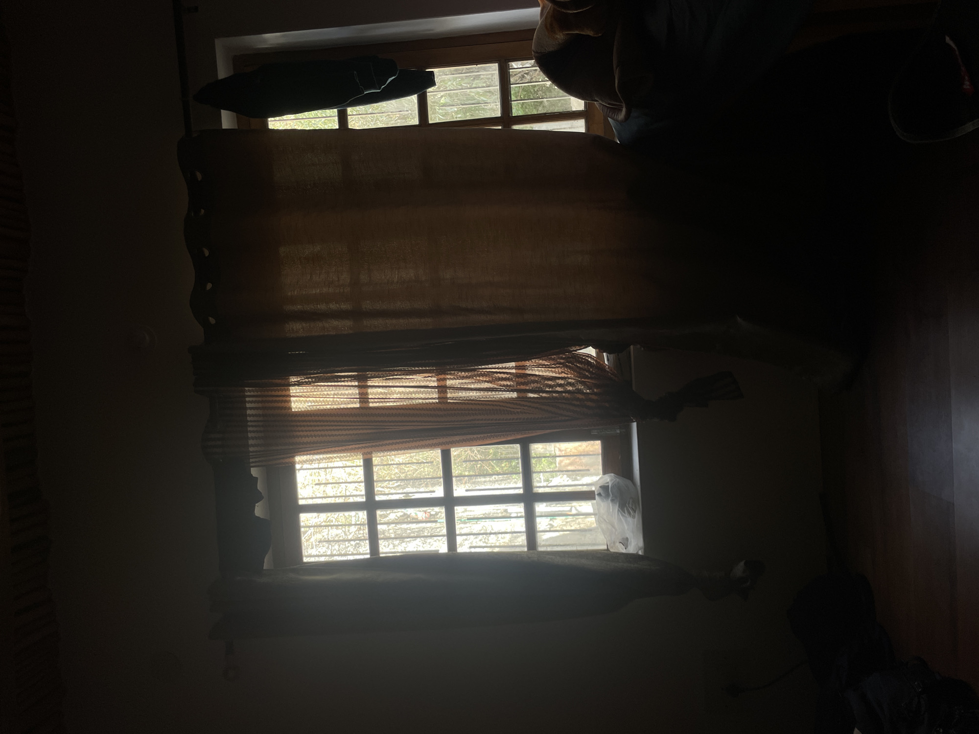 The image depicts an interior scene, possibly a room, with two windows covered by curtains. The light filtering through the windows suggests daytime. The left window is partially obscured by a sheer, patterned curtain, revealing a barred windowpane and a white plastic bag on the windowsill. The right window is covered by a heavier, opaque curtain, with a dark object hanging on the outside of the window. The room appears to be sparsely furnished, with some items of clothing or bags visible on the floor in the lower left and right corners. The overall lighting is dim, with strong contrasts between the illuminated windows and the shadowed interior. The context of Leh, India is not visually apparent from the image.