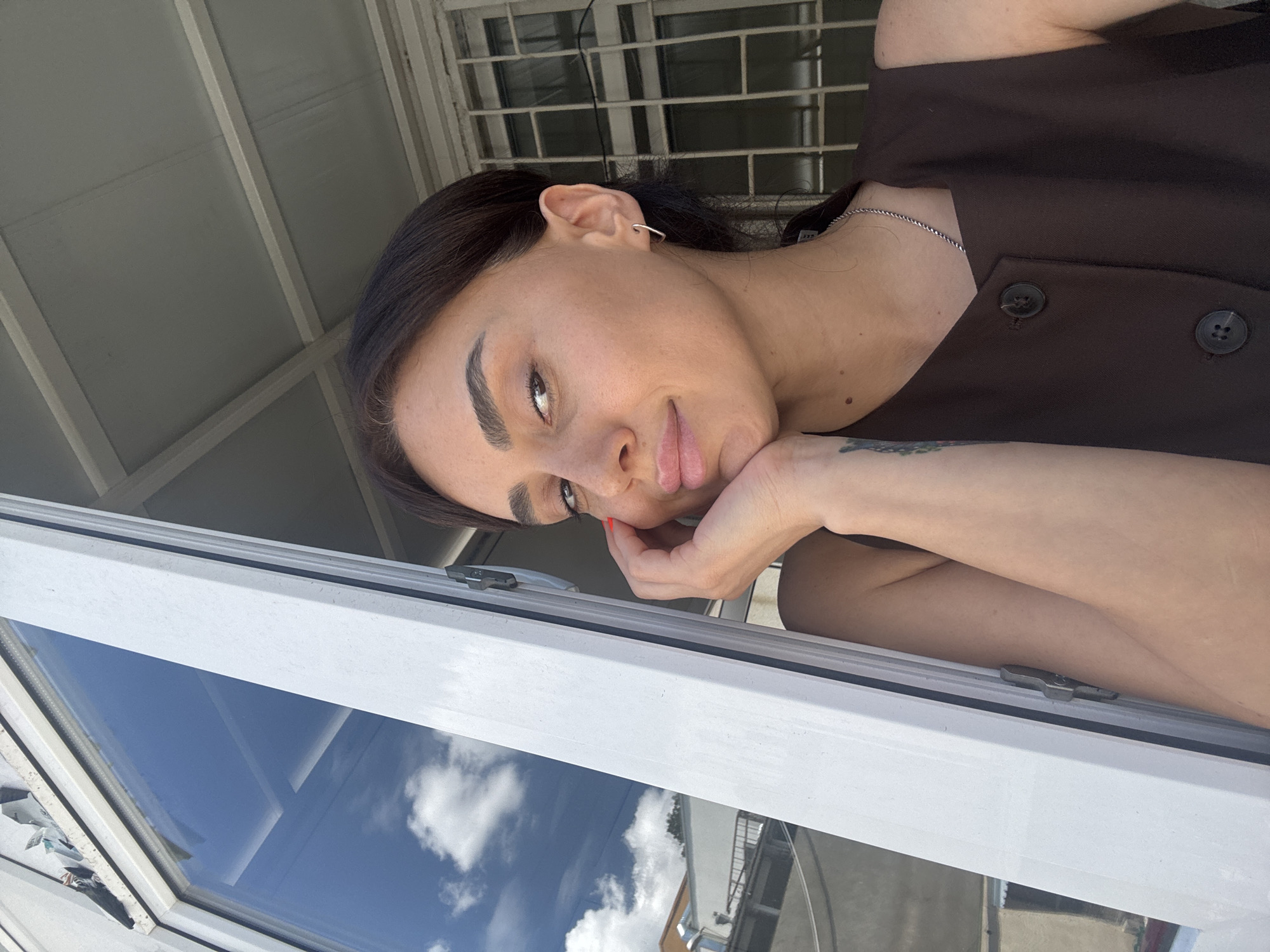 A woman is captured in a close-up selfie, leaning her head on her hand as she looks at the camera. She has dark hair pulled back, and is wearing a sleeveless dark brown top with two visible buttons. Her left ear has a small silver hoop earring. She is positioned near an open white window frame, through which a bright blue sky with white clouds is visible. Reflected in the window are buildings and trees. The background appears to be an urban setting with a barred window on the right side. The lighting suggests it is daytime, with sunlight casting shadows and highlighting features. The image is taken from a slightly elevated perspective.