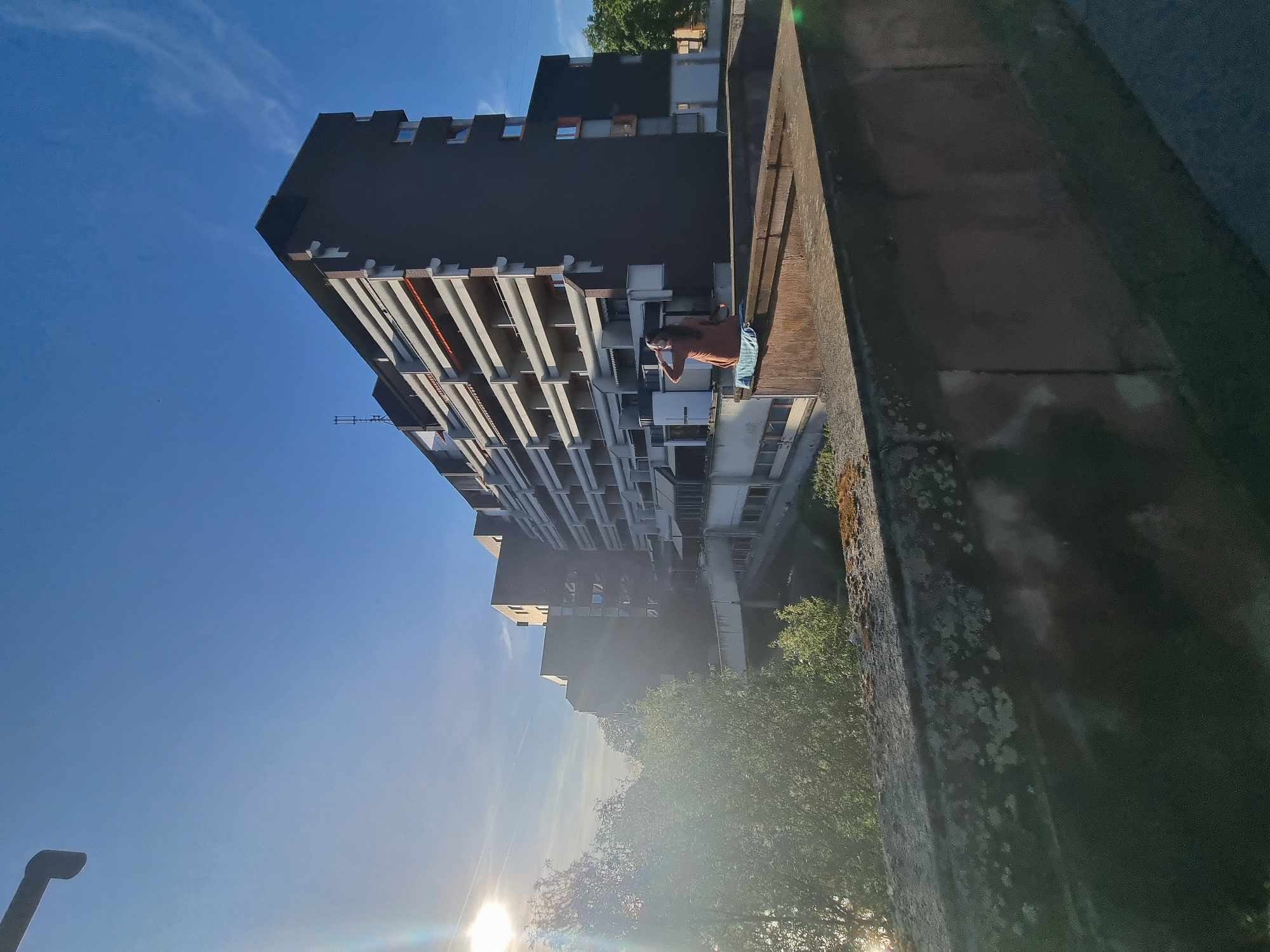 The image shows a tall, modern building with multiple balconies. A person is standing on one of the balconies, looking towards the street. The building is located in the city of Saarbrücken, Germany. The sky is clear and blue with the sun shining brightly. Trees are visible in the foreground, suggesting the building is in a relatively green area. A concrete structure is in the foreground, acting as a partial barrier.