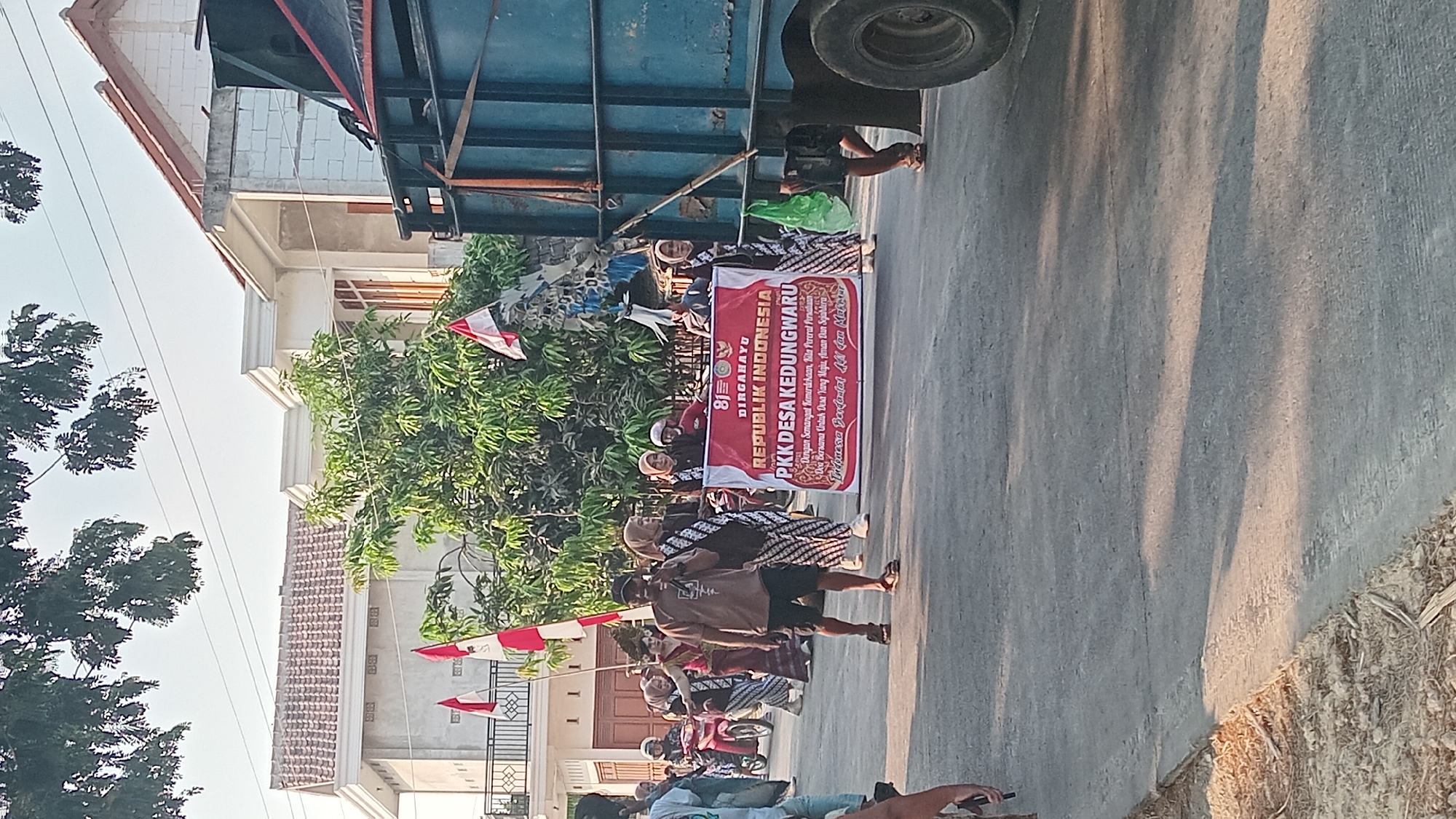 The image depicts a street scene in Kedungwaru, Indonesia, likely during a procession or event. A large banner held by several individuals reads "81 DIRGAHAYU REPUBLIK INDONESIA PKK DESA KEDUNGWARU" and below that, "Dengan Semangat Kemerdekaan, Kita Pererat Persatuan. Do'a Bersama Untuk Desa Yang Maju, Aman Dan Sejahtera. Terus Berfastat Aat Dan Makmur". The banner is predominantly red and white.
Several people are walking in the street, some carrying Indonesian flags. One man in the foreground is speaking into a microphone, wearing a brown t-shirt and shorts. Women in the procession are wearing traditional clothing with batik patterns and headscarves. Behind the procession, a large blue truck is visible, partially obscuring the view of buildings. The environment suggests a residential area with houses visible in the background, some under construction. The lighting indicates it is daytime, with sunlight casting shadows on the road. The ground is paved, and there are trees lining the street. The overall impression is a community gathering or celebration.
