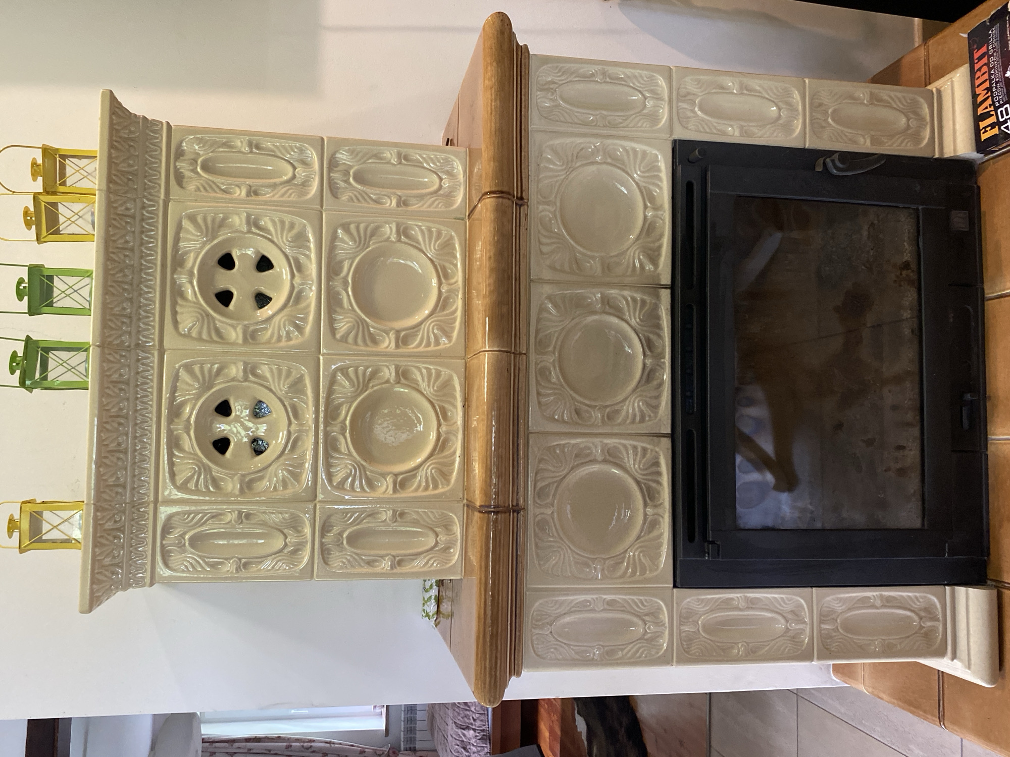 The image shows a fireplace with decorative ceramic tiles, located in Biała Wieś, Poland. The fireplace has a wooden mantel and a black metal insert with a glass door. The ceramic tiles are cream-colored and feature embossed floral and geometric patterns. On top of the fireplace, there are several small decorative lanterns. The background suggests an indoor setting, possibly a residential interior, with part of a wall and a window visible on the left. A box of fire starters labeled "FLAMBIT" is placed on the wooden surface to the right of the fireplace. The lighting is natural, coming from the window and ambient room light.